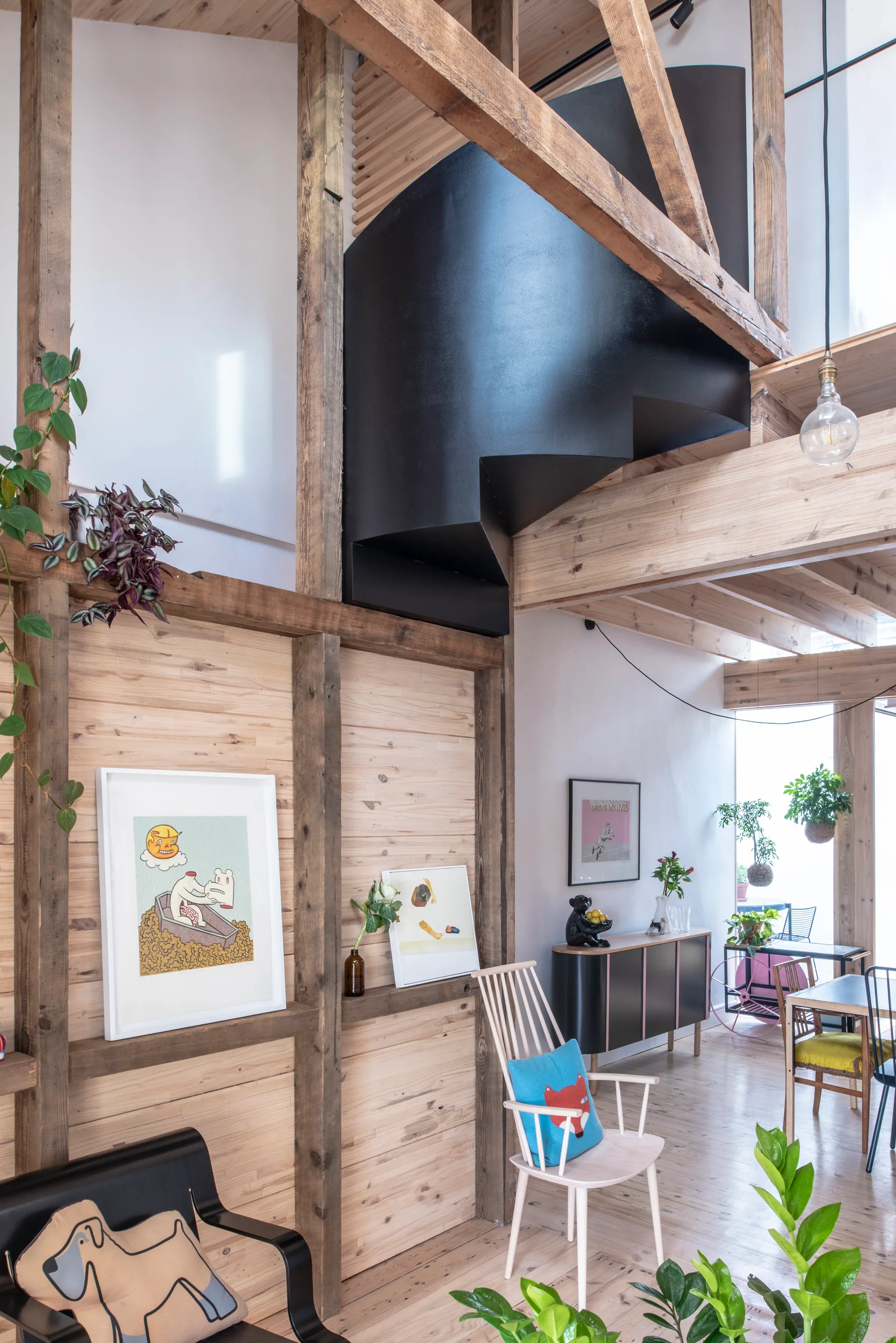 An open living space features exposed timber framing, a sculptural black staircase, light wood-slatted walls, and contemporary chairs.