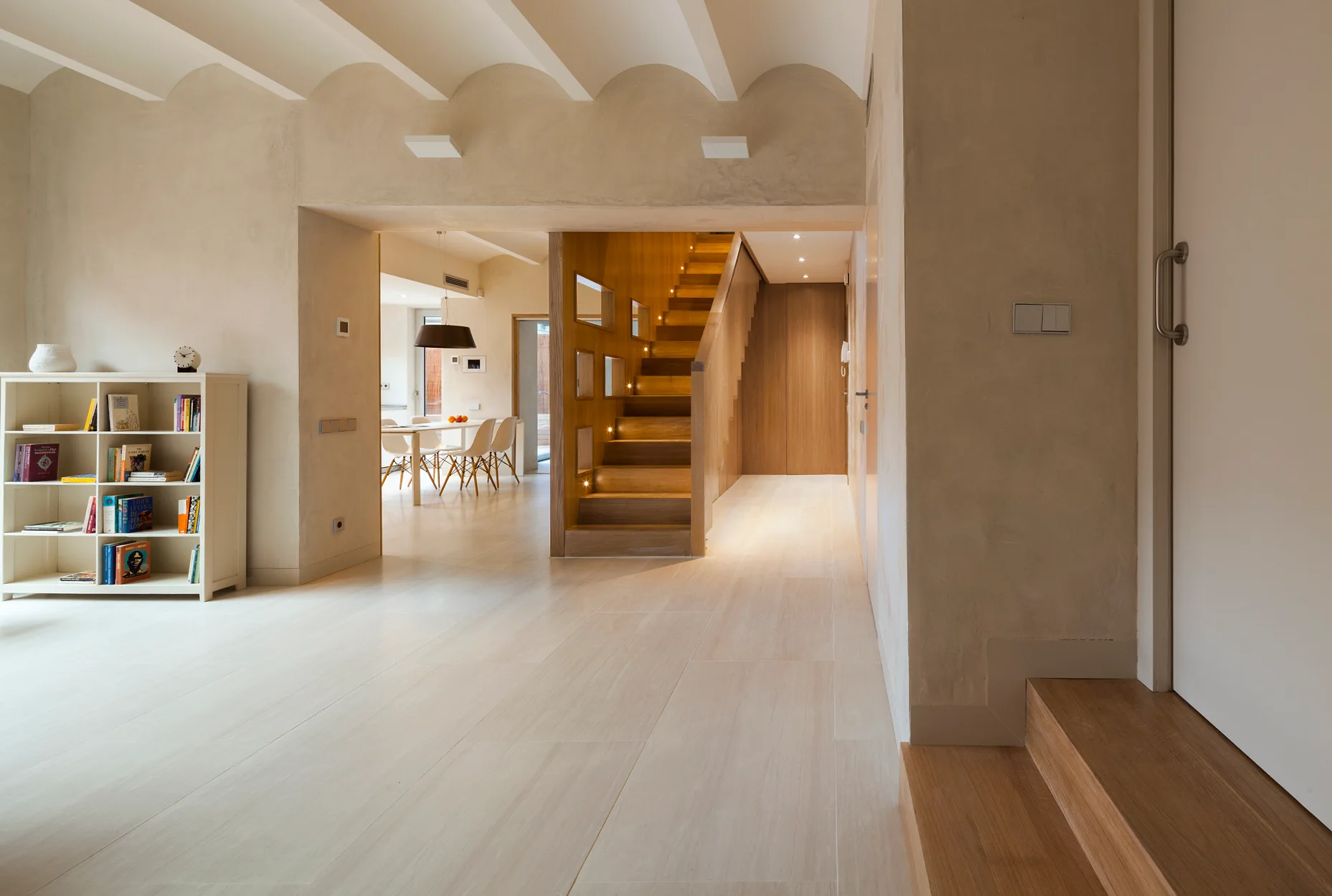 An open home interior features a central wooden staircase with integrated lights, matte plaster walls, and a low white bookshelf.