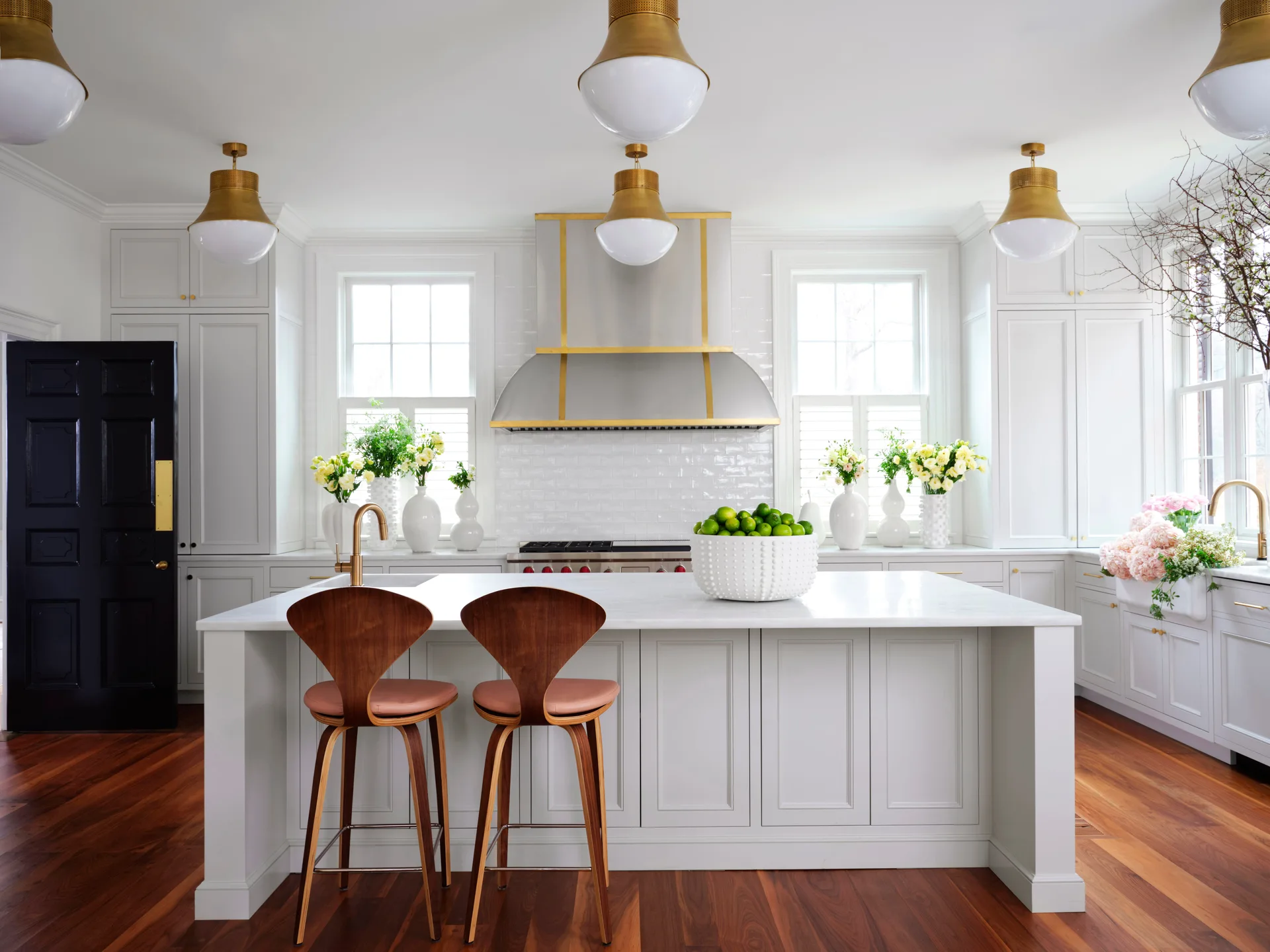 A symmetrical white kitchen with a marble-topped island, curved wood bar stools, a brass-trimmed range hood, and gold pendant lights.