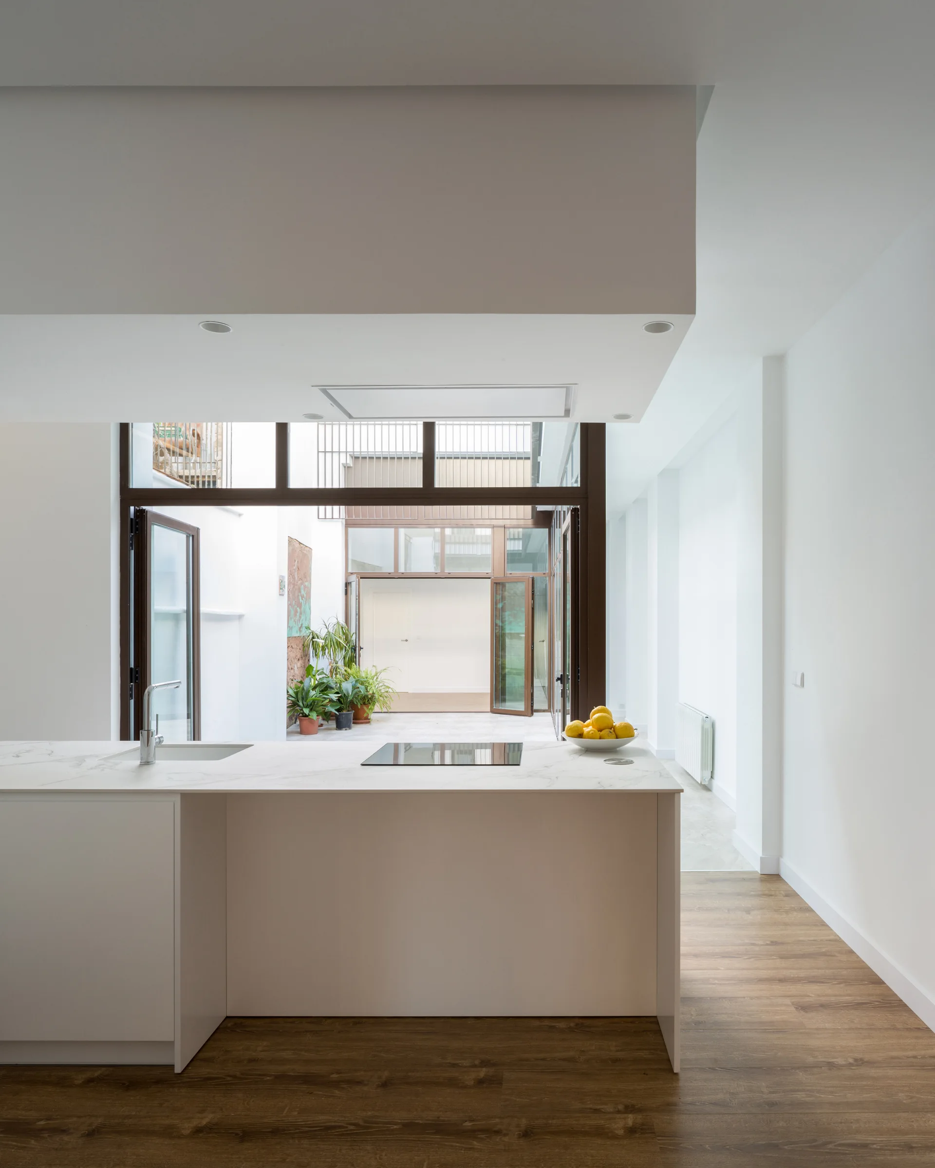 A minimalist white kitchen island with a stone-look countertop stands on wood floors in front of large glass doors opening to an outdoor courtyard.
