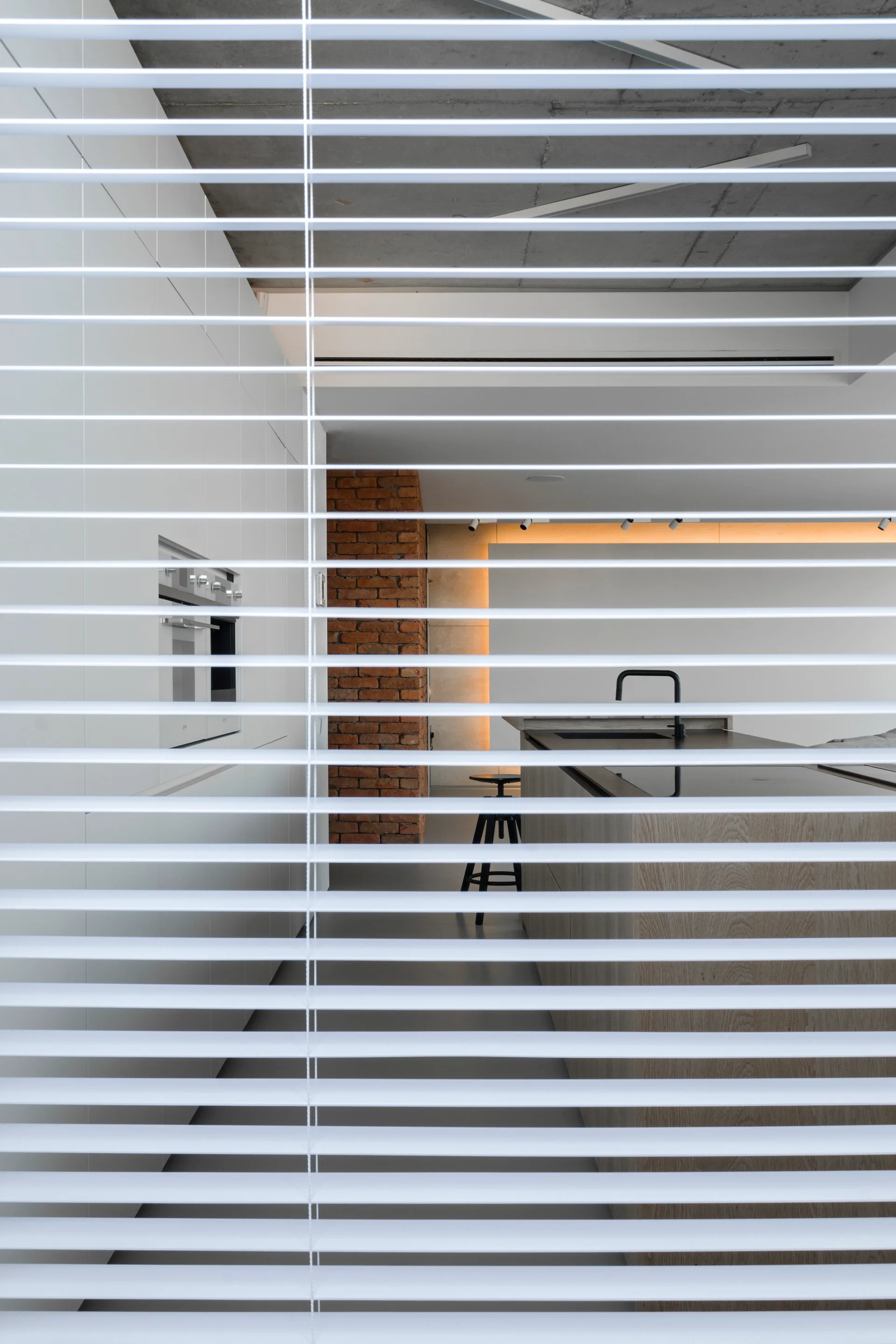 A modern kitchen with a wood island and brick column is viewed through the horizontal slats of white window blinds.