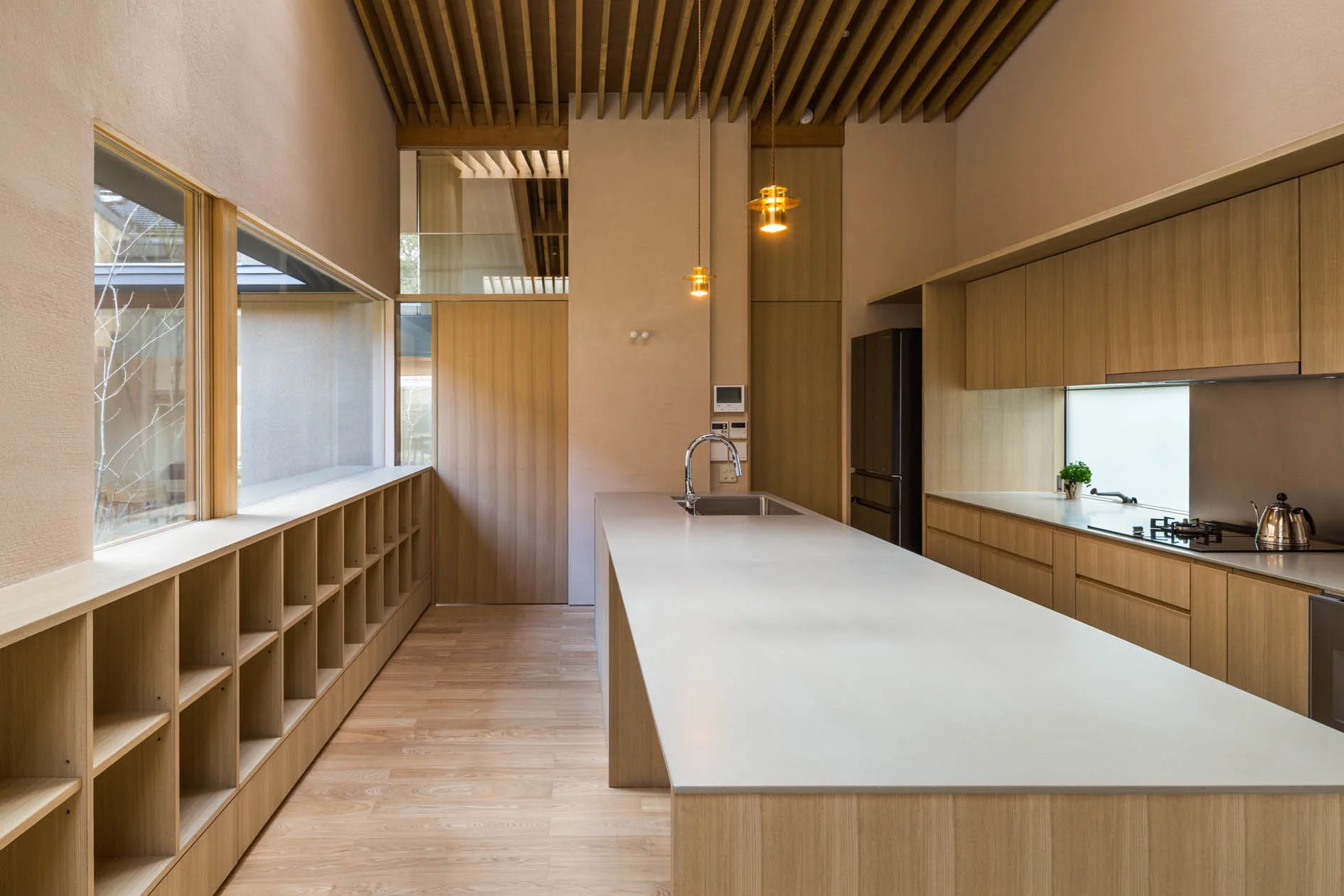 A modern minimalist kitchen features a long white island, light wood cabinetry, a slatted ceiling, and low built-in wooden shelving under a horizontal window.