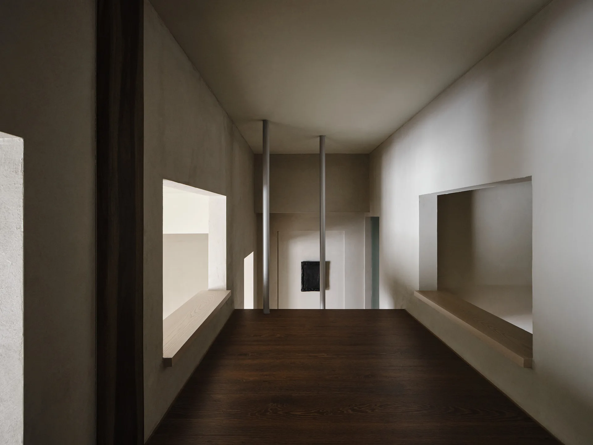 A minimalist hallway features plaster walls with rectangular cutouts, dark wood flooring, and two slim metal columns leading to a lower level.