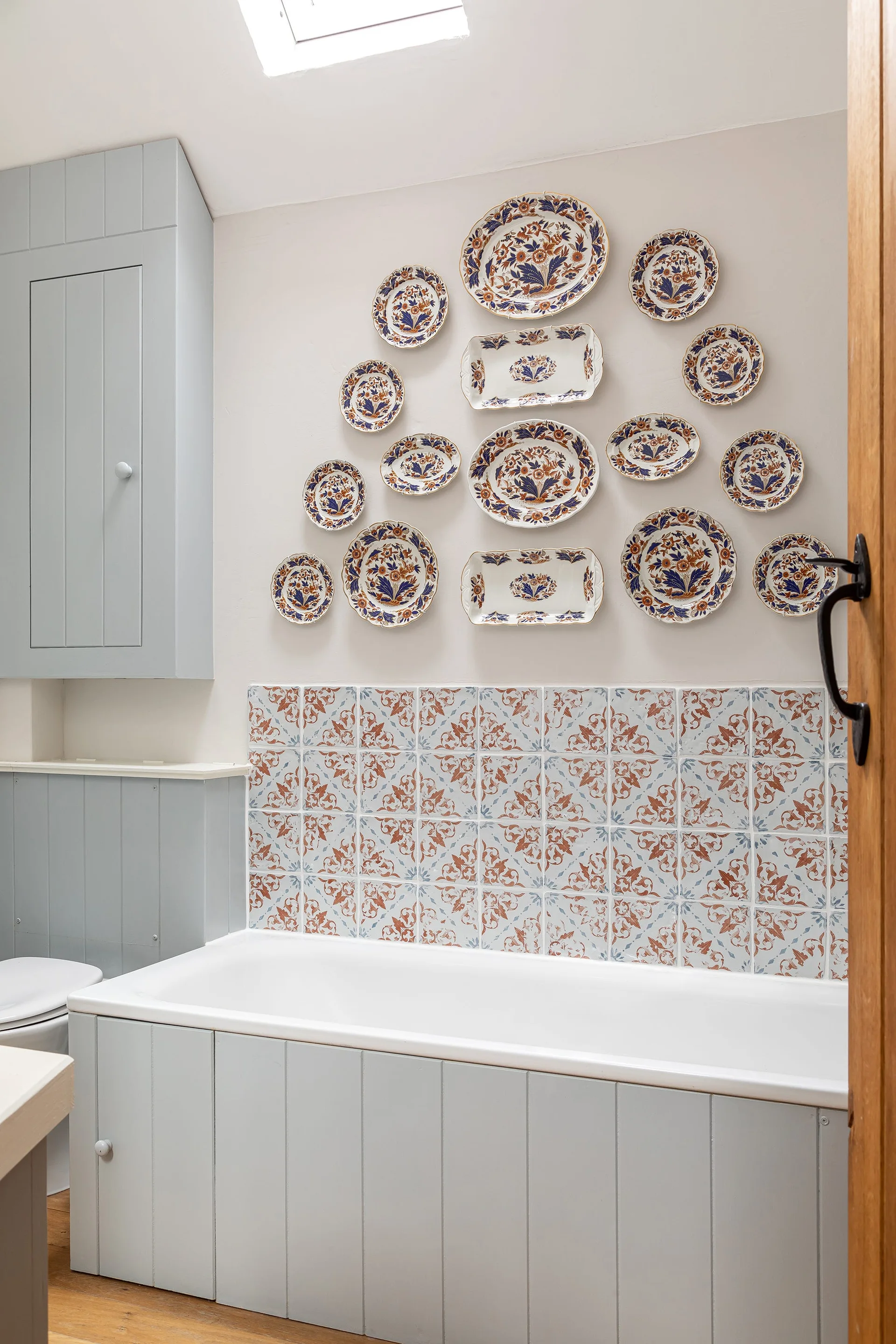 A light blue beadboard-clad bathtub sits below a floral patterned tile backsplash and an artistic arrangement of ceramic plates on an off-white wall.