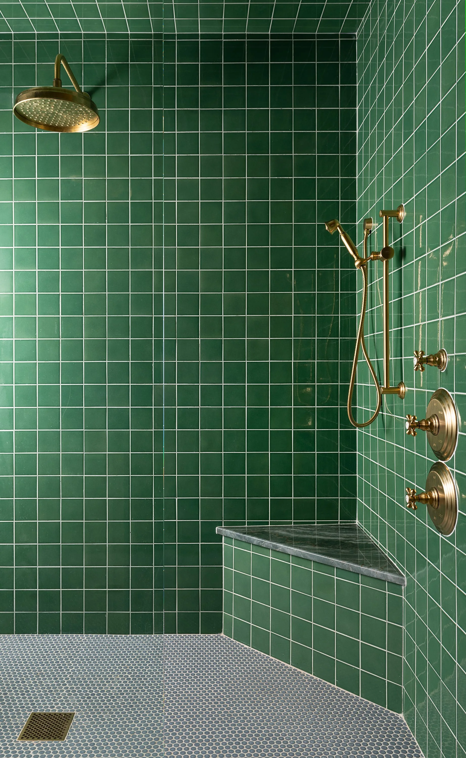 A shower enclosure features glossy green square wall tiles, brass hardware, a grey penny-tile floor, and a built-in corner bench with a grey stone top.