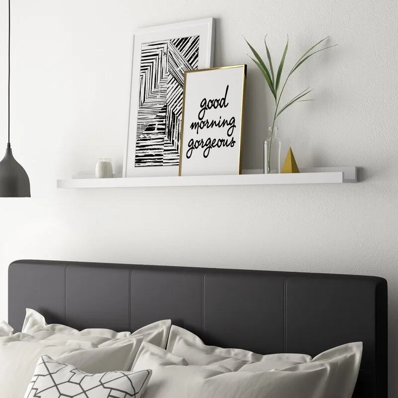 A grey upholstered headboard is positioned beneath a white floating shelf decorated with framed art, a small plant, and a gold accent piece.