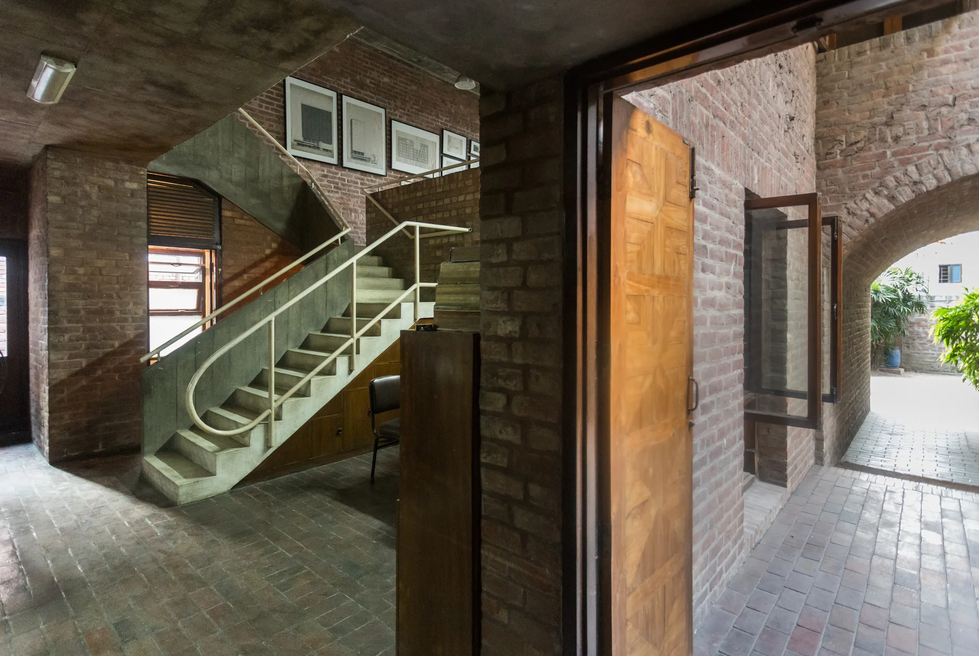 A concrete staircase with a metal railing stands in an entryway featuring exposed brick walls, brick tile floors, and an open wooden door.
