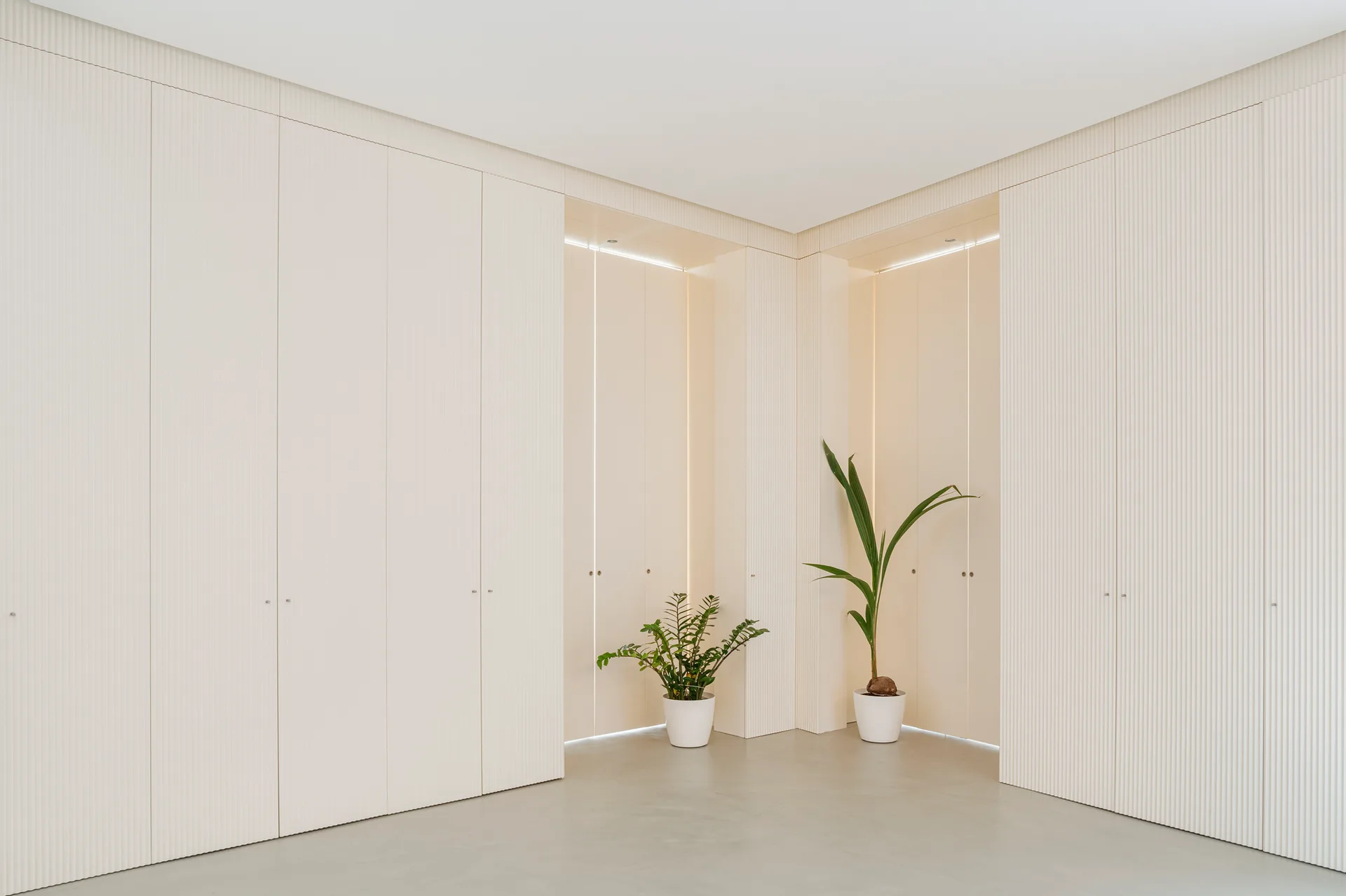 Minimalist off-white hallway featuring floor-to-ceiling fluted cabinets, backlit niche openings, and two green potted plants on a seamless grey floor.