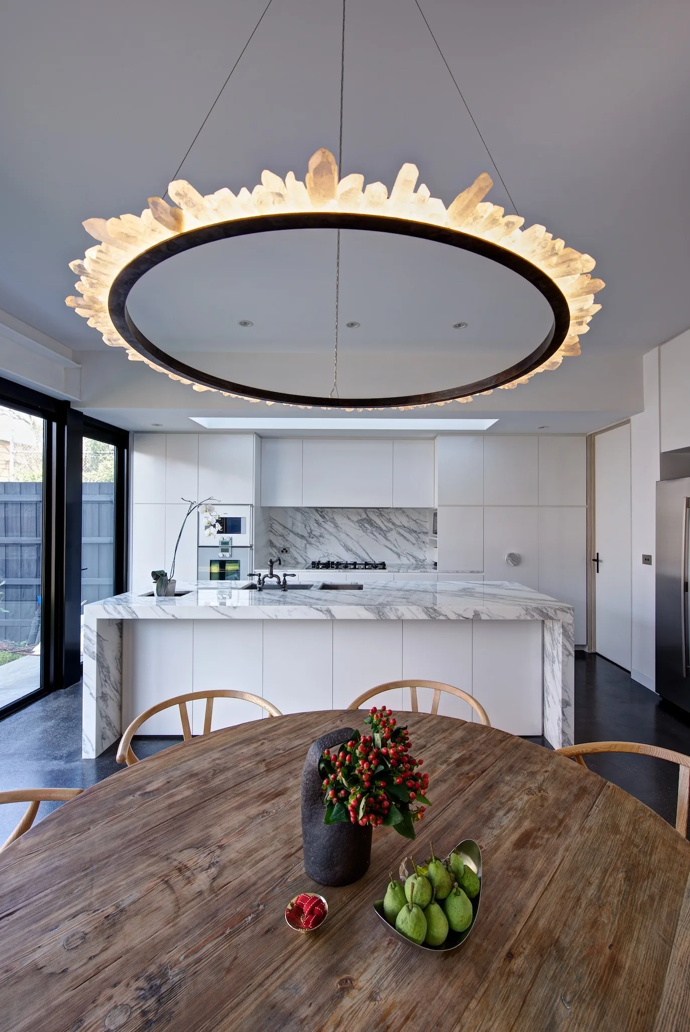 A modern kitchen features a marble waterfall island, flat-panel white cabinets, a round wooden dining table, and a large circular crystal chandelier.