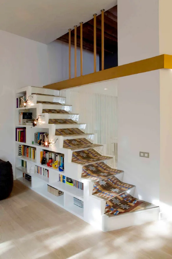 A white staircase features an integrated bookshelf built into its side and colorful geometric patterned tile designs on each tread.