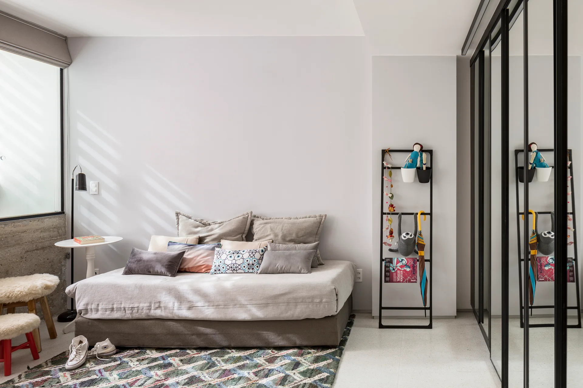 A minimalist bedroom features a low daybed with grey pillows, a patterned rug, a black leaning ladder shelf, and a large mirrored wardrobe.