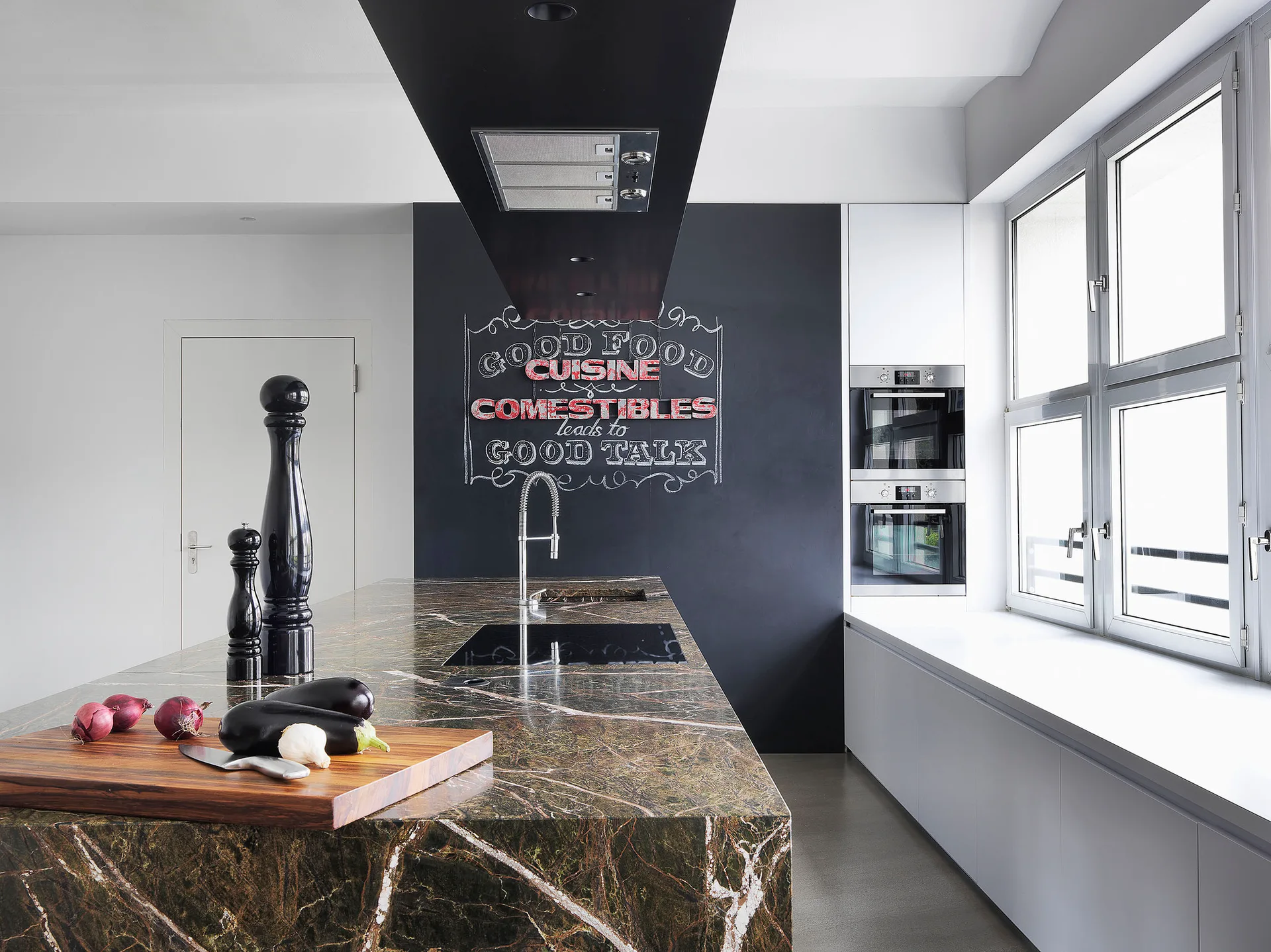 A modern kitchen featuring a dark veined stone island, a white perimeter counter beneath casement windows, and a black chalkboard wall.