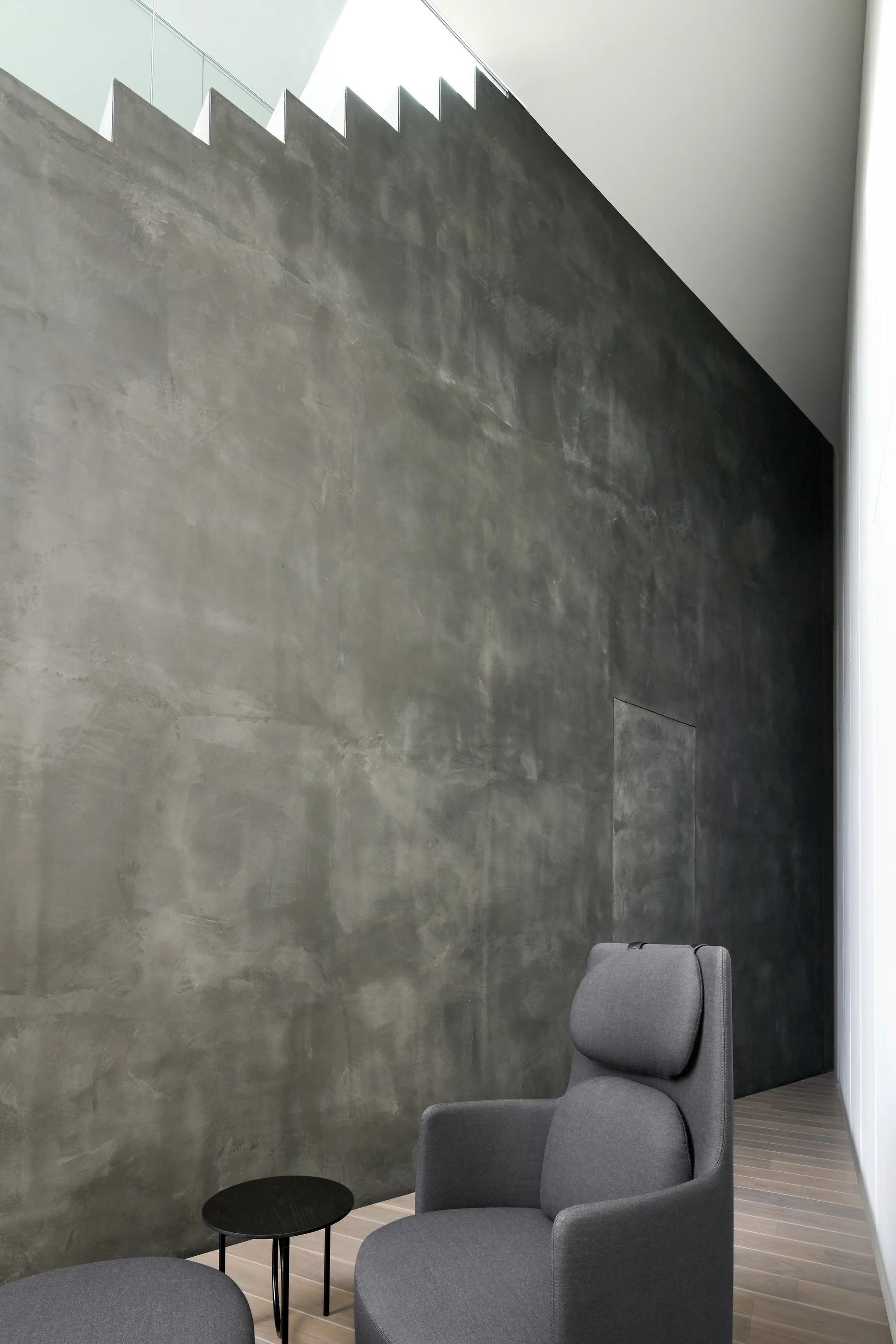 A minimalist grey armchair and small black side table sit in front of a tall textured concrete wall beneath a jagged staircase silhouette.