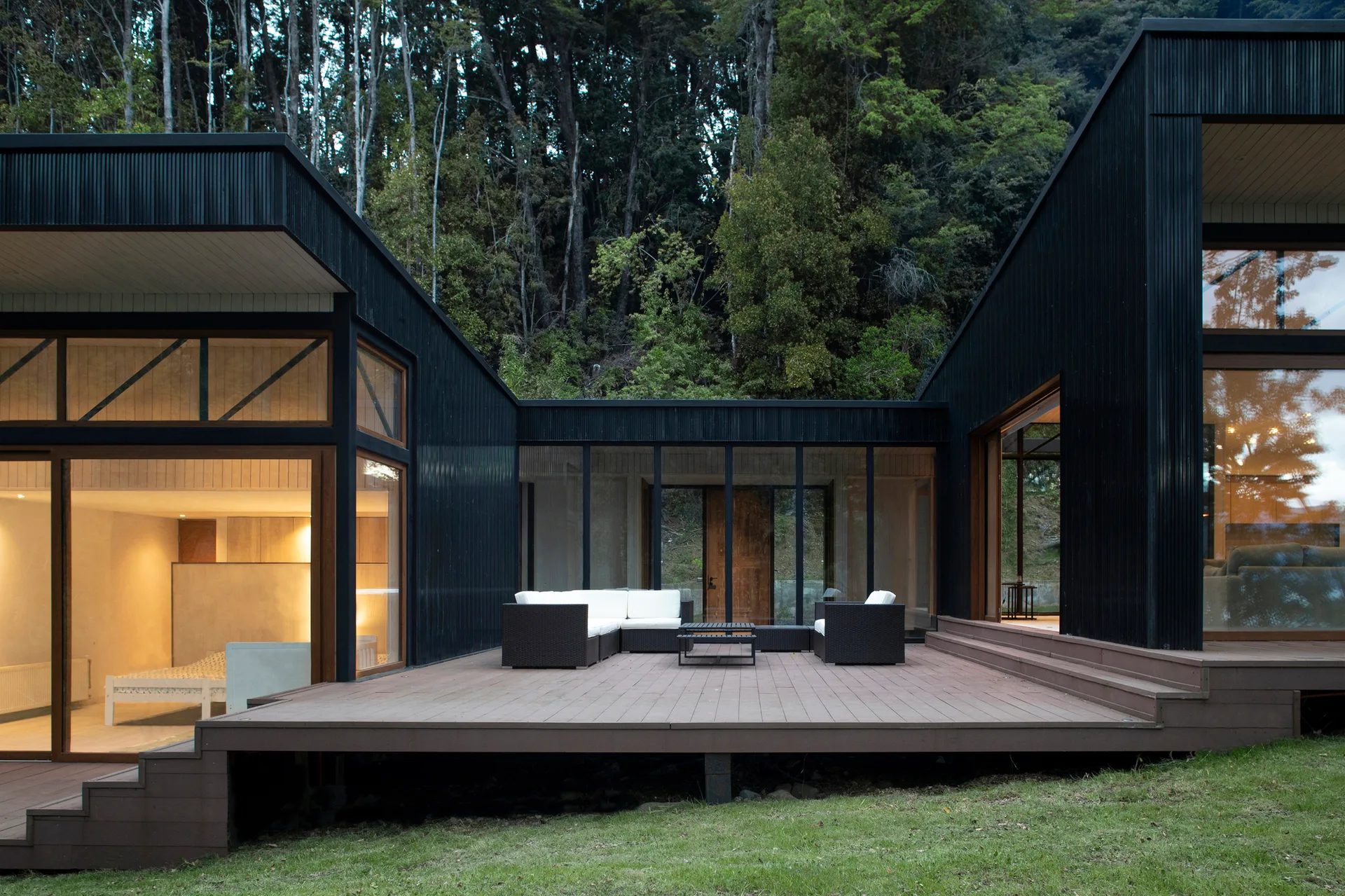 A modern black-clad house features an expansive wooden deck with a white-cushioned wicker sofa set, backed by a dense green forest.