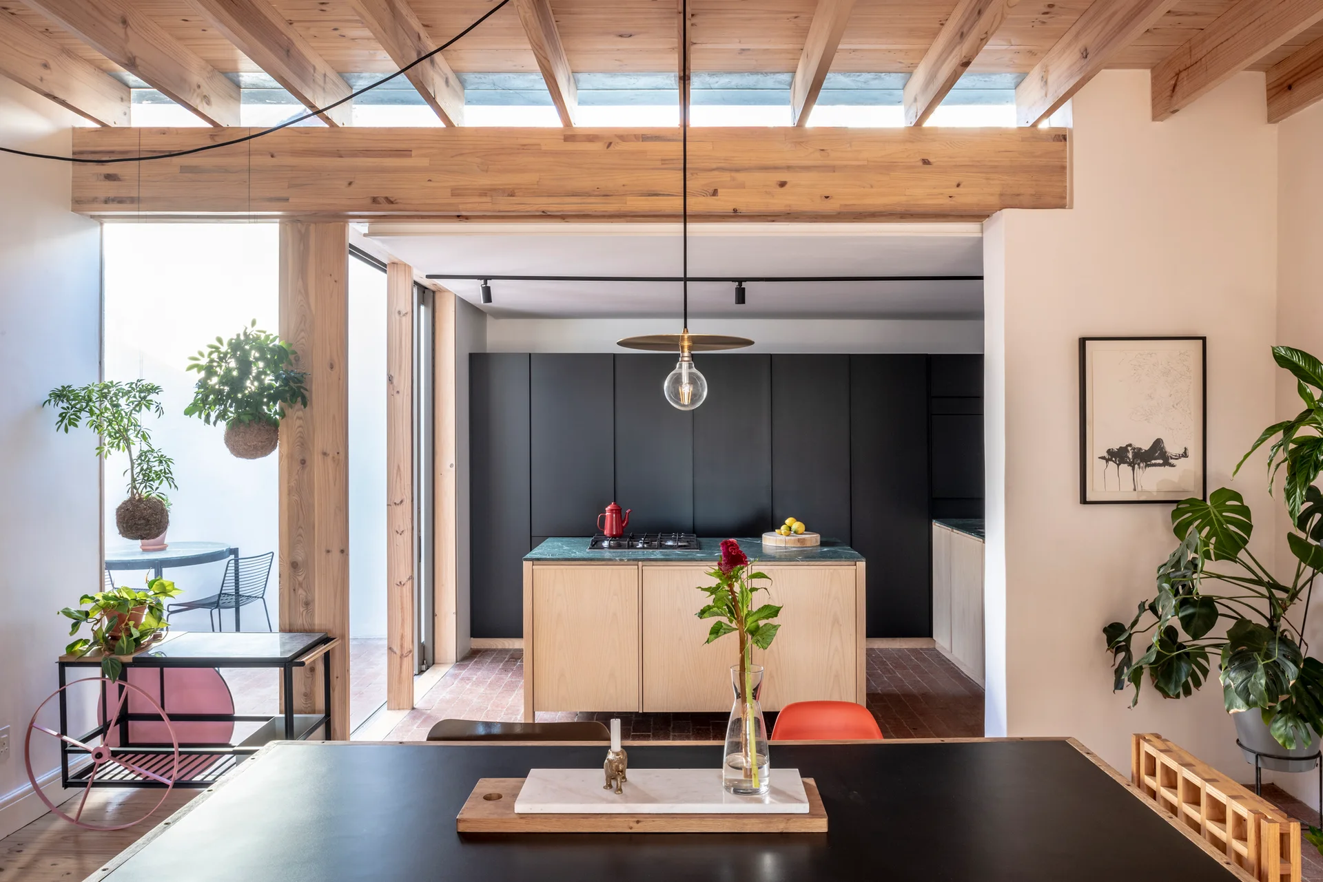A modern kitchen and dining space featuring a light wood island with a green stone countertop, matte black cabinets, and exposed ceiling beams.