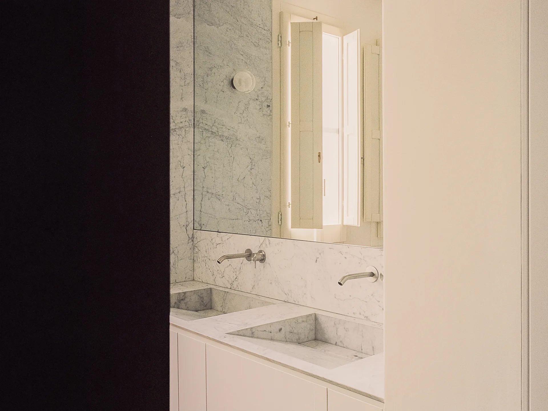 A minimalist bathroom vanity features two integrated marble basins, wall-mounted metal faucets, and a large mirror reflecting wooden window shutters.