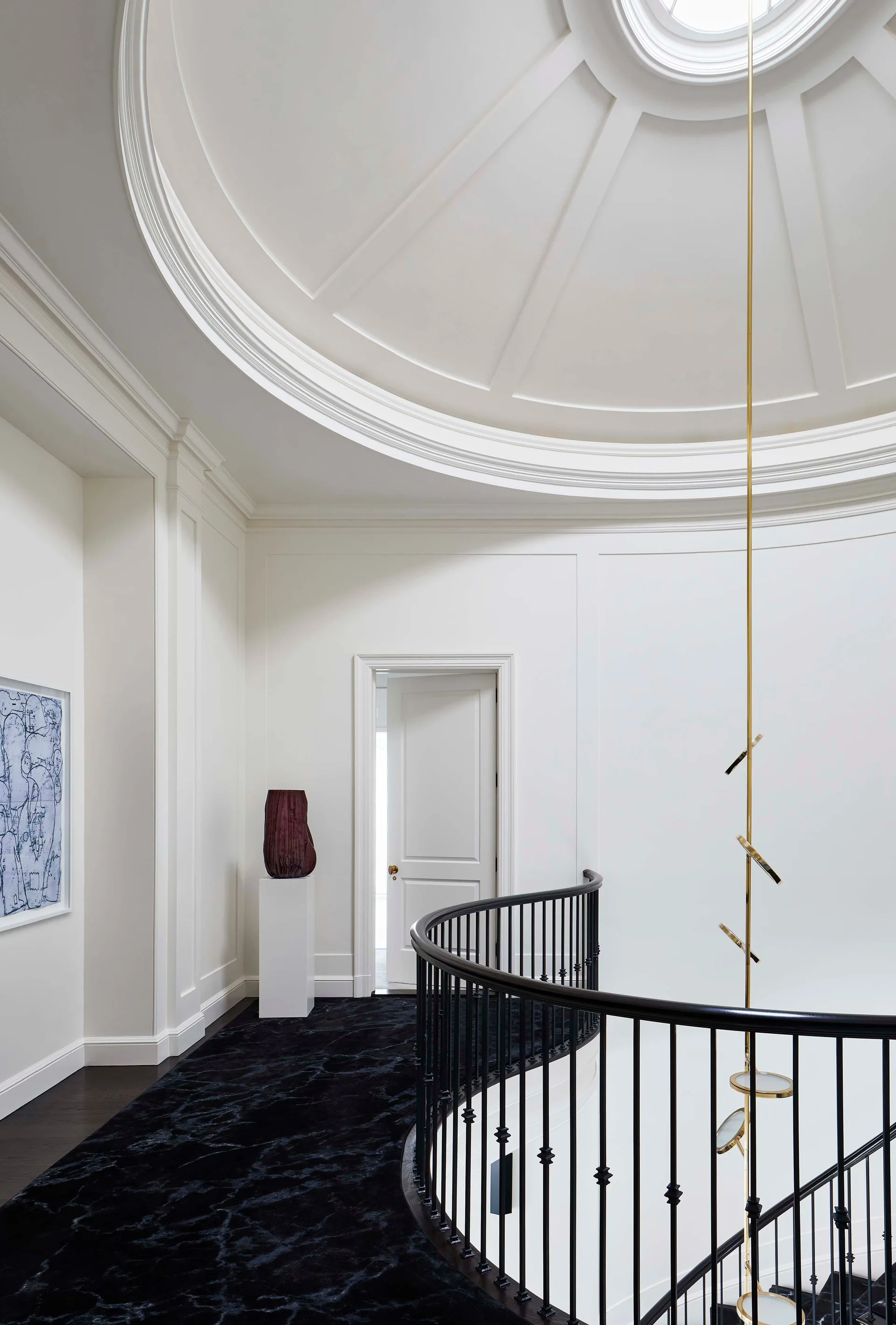 A curved black staircase railing wraps around a landing with white paneled walls, a domed ceiling, and a vertical gold pendant light.