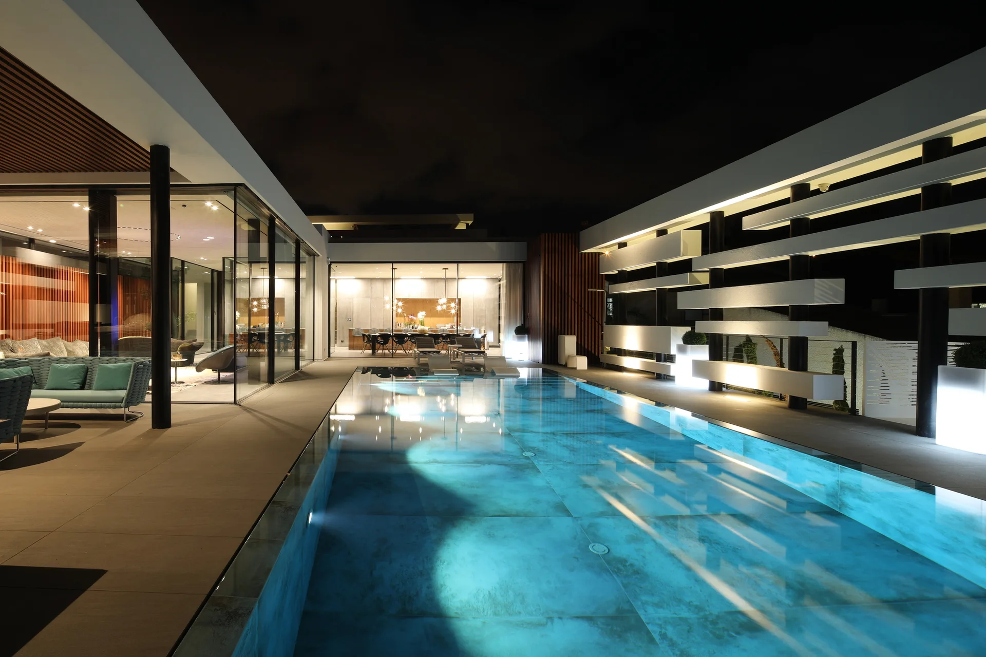An illuminated contemporary swimming pool at night is bordered by large-format grey patio tiles, glass walls, and a white architectural screen wall.
