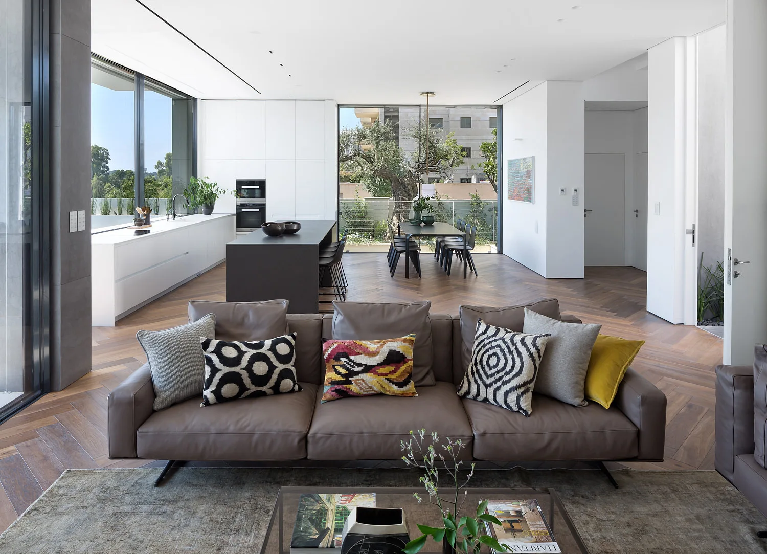 A contemporary open-plan living room and kitchen featuring a brown leather sofa, herringbone wood floors, and a dark kitchen island.