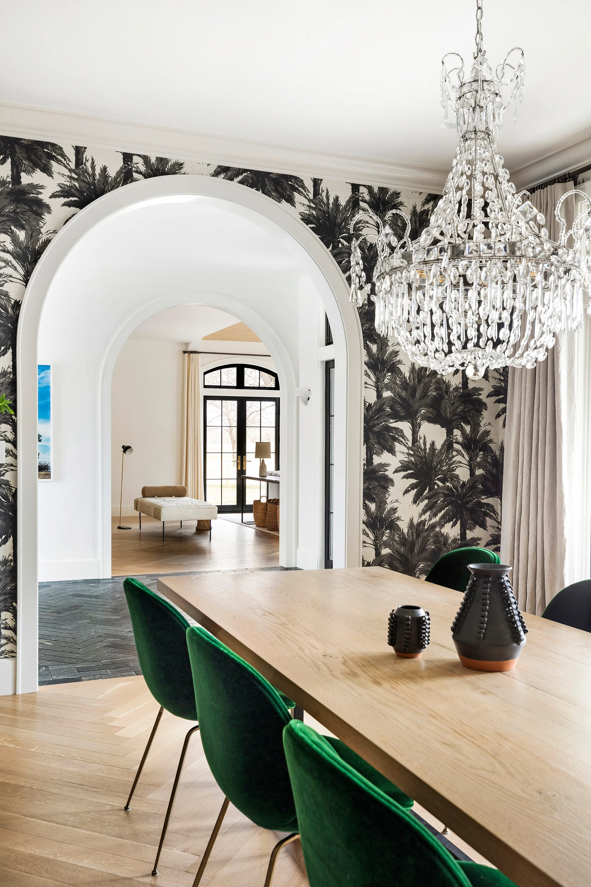 A dining room features green velvet chairs around a wood table beneath a crystal chandelier, with palm-patterned wallpaper framing a large white archway.