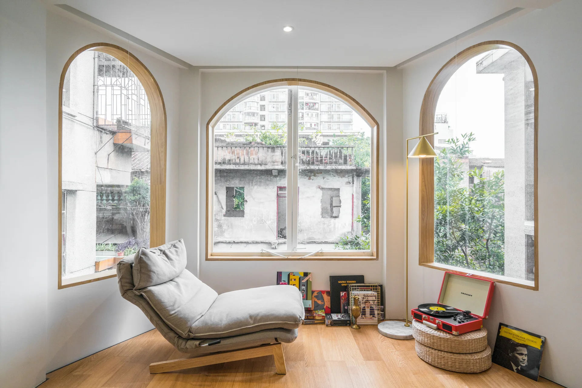A grey lounge chair rests near three large arched windows with wooden trim in a minimalist room with wood floors and a vintage record player.