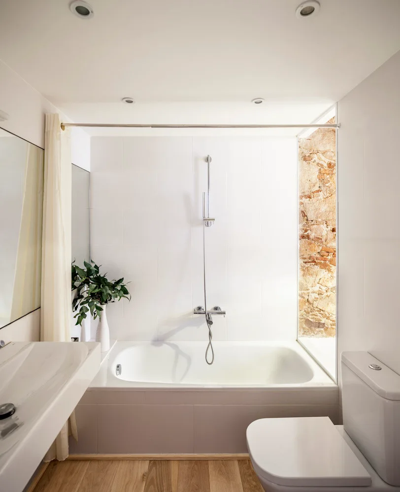 A minimalist white bathroom featuring a built-in bathtub, light wood floors, and a vertical recess of exposed raw stone.