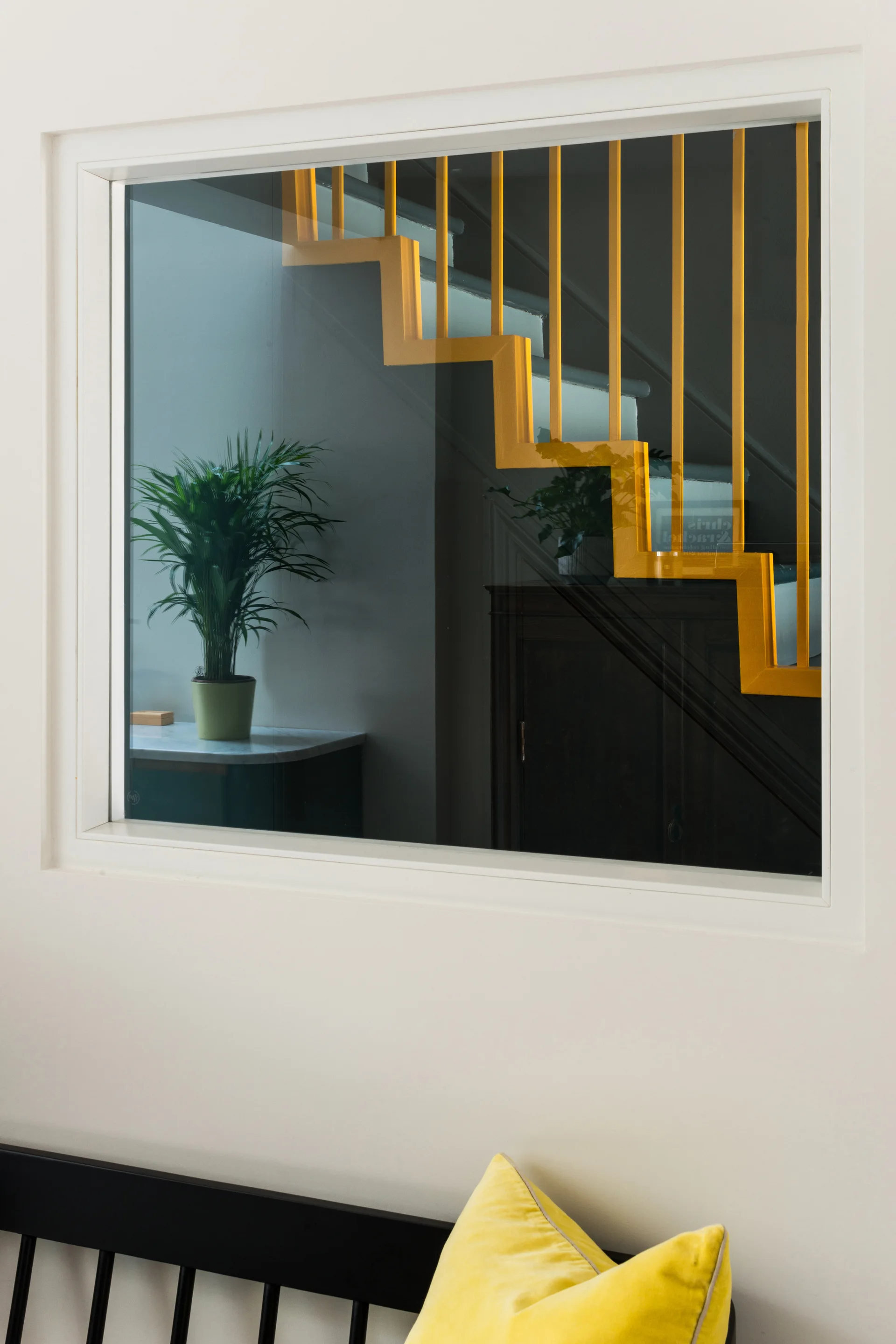 A view through a white-framed interior window showing a staircase with a bright yellow railing, a potted plant, and a black bench with a yellow pillow.