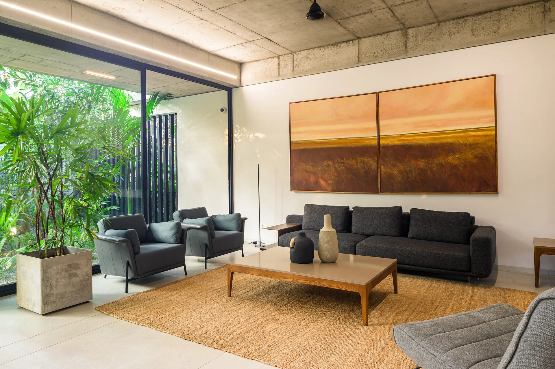 A charcoal sofa and two matching armchairs sit on a textured woven rug beneath an exposed concrete ceiling and a large landscape diptych painting.