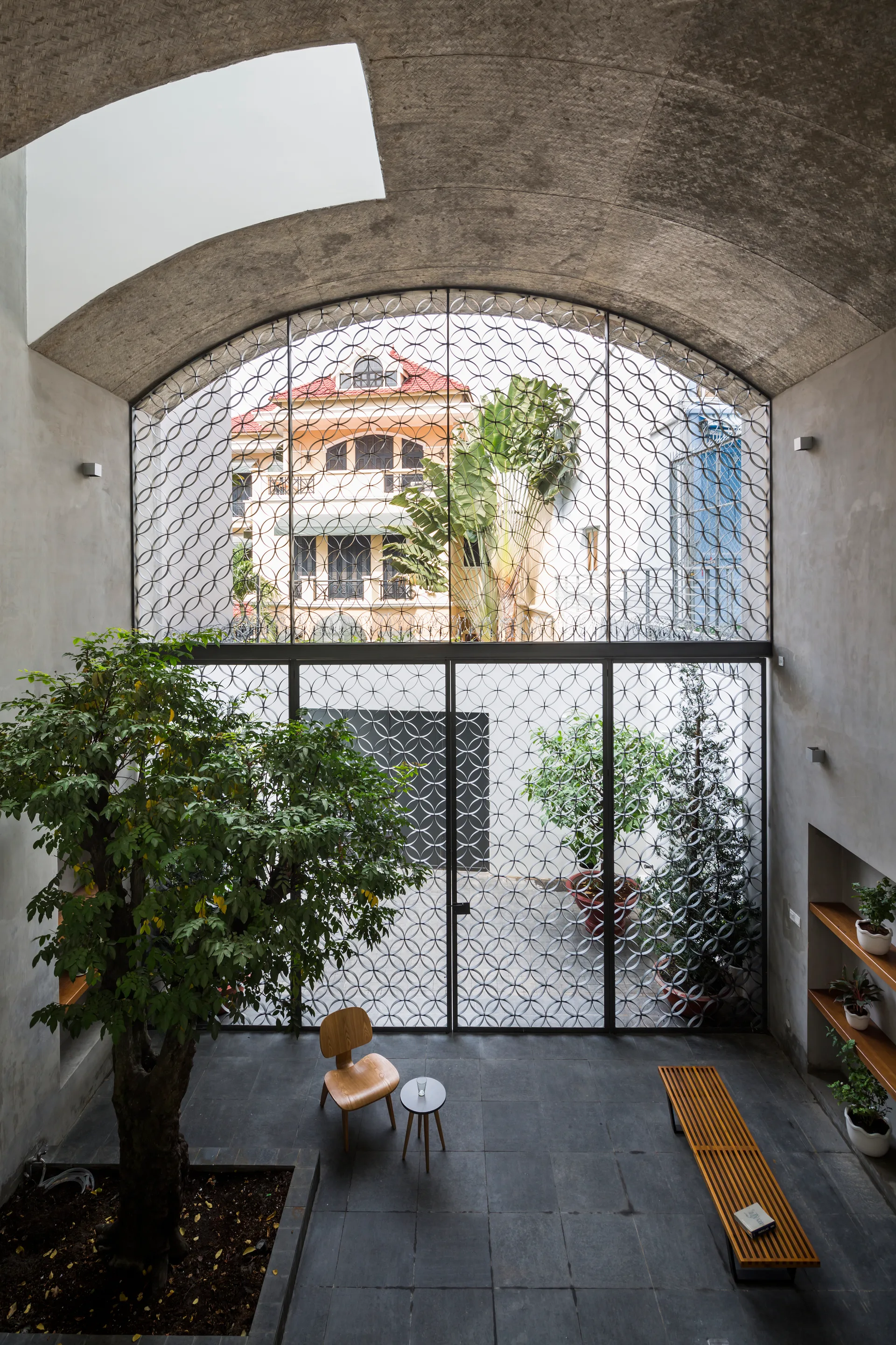 A minimalist interior atrium features a tall arched metal screen, a tree growing from the stone floor, concrete walls, and simple wood furniture.