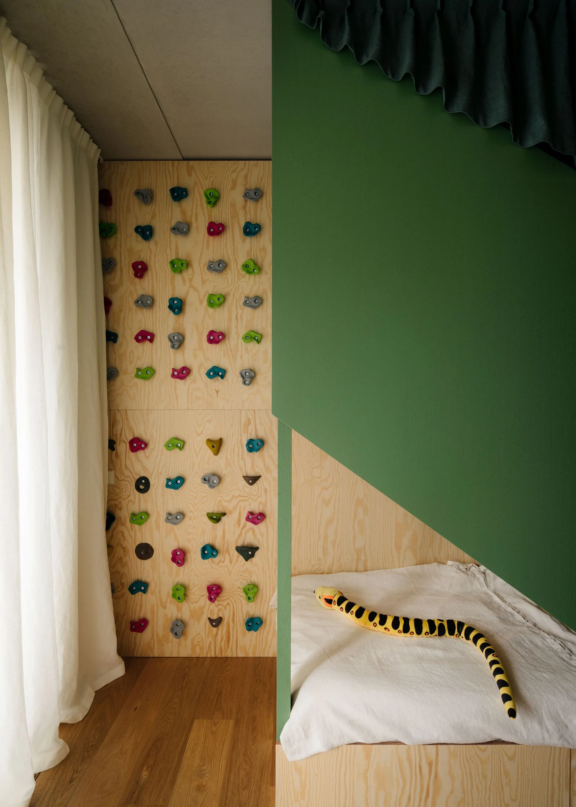A children's room with a plywood climbing wall featuring colorful handholds next to a built-in wooden bed with a green partition.