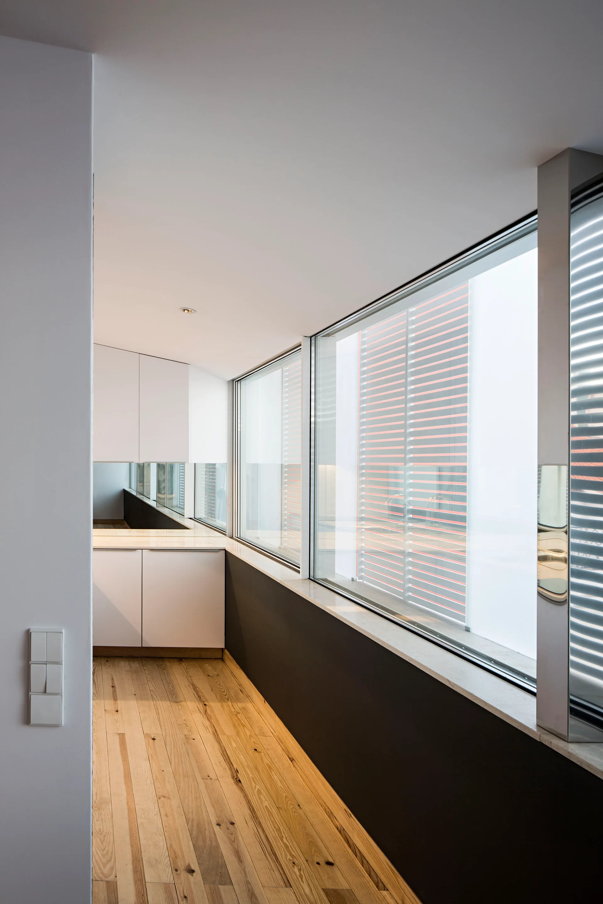 A minimalist corridor kitchen features light wood floors, white cabinetry, and a long sequence of large windows above a dark gray wall.