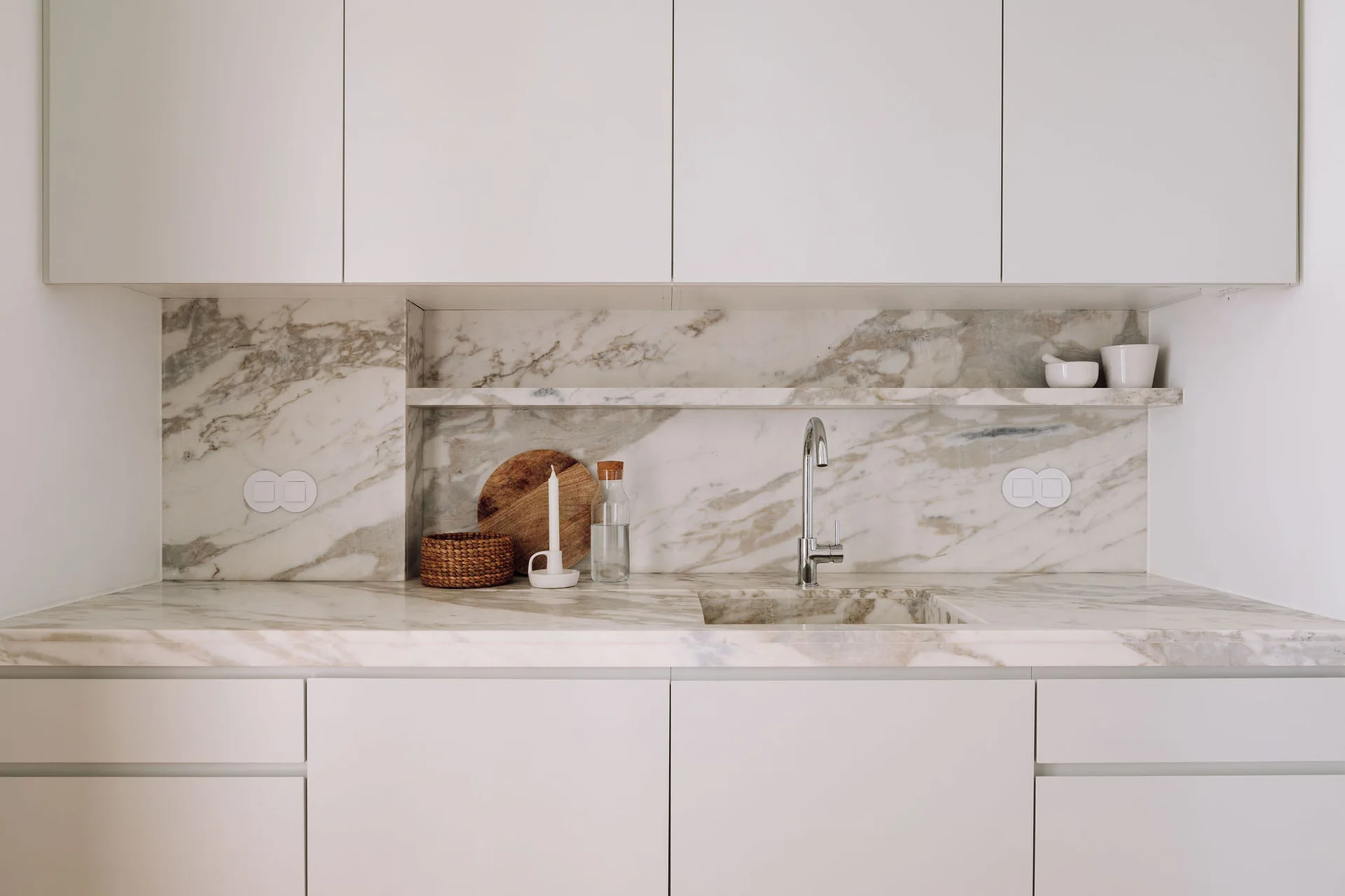 Minimalist white cabinetry paired with a heavily veined marble countertop, an integrated sink, and a matching stone backsplash with a shelf.