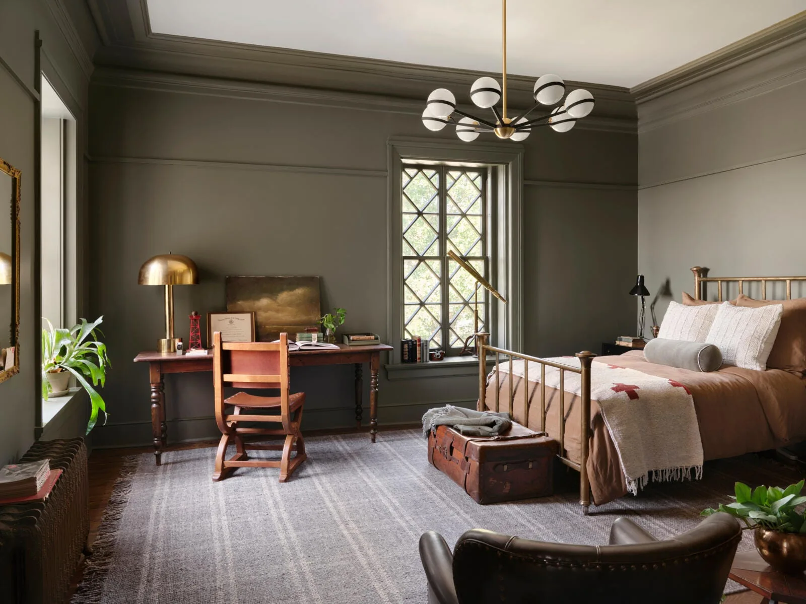 A moody bedroom features olive green walls, a brass metal bed frame, a vintage wooden desk with a leather chair, and a plaid rug.