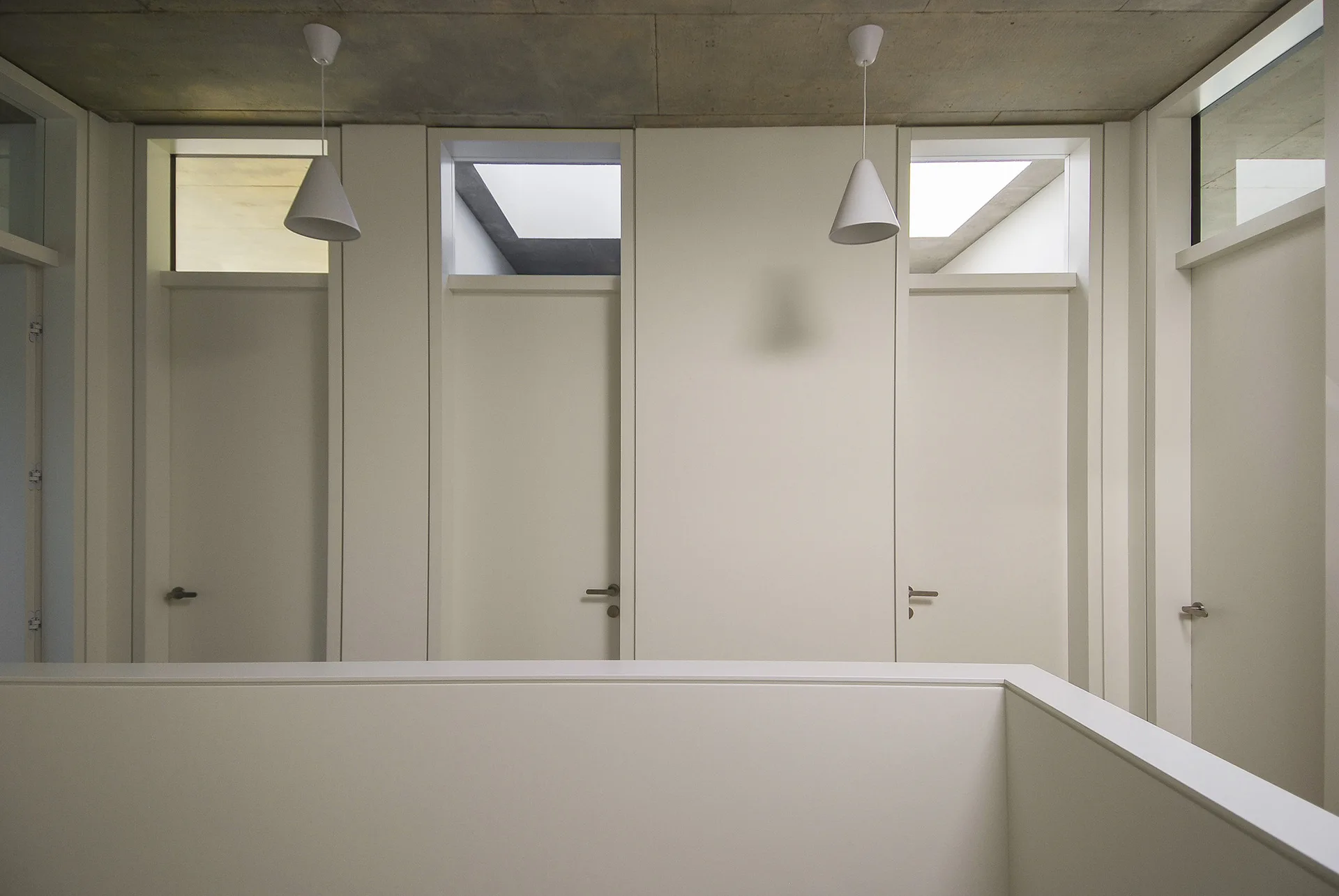 A minimalist hallway with white doors topped by small transom windows beneath a raw grey concrete ceiling with two white pendant lights.