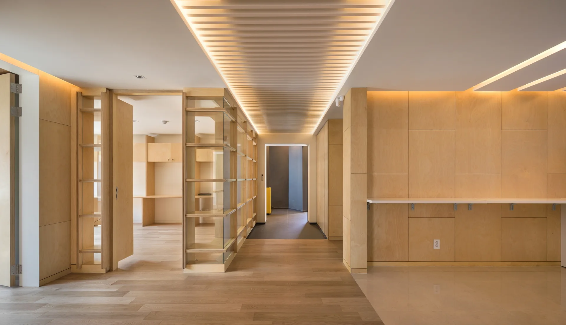 A minimalist modern corridor with light wood wall paneling, a slatted wood ceiling, and warm indirect lighting.