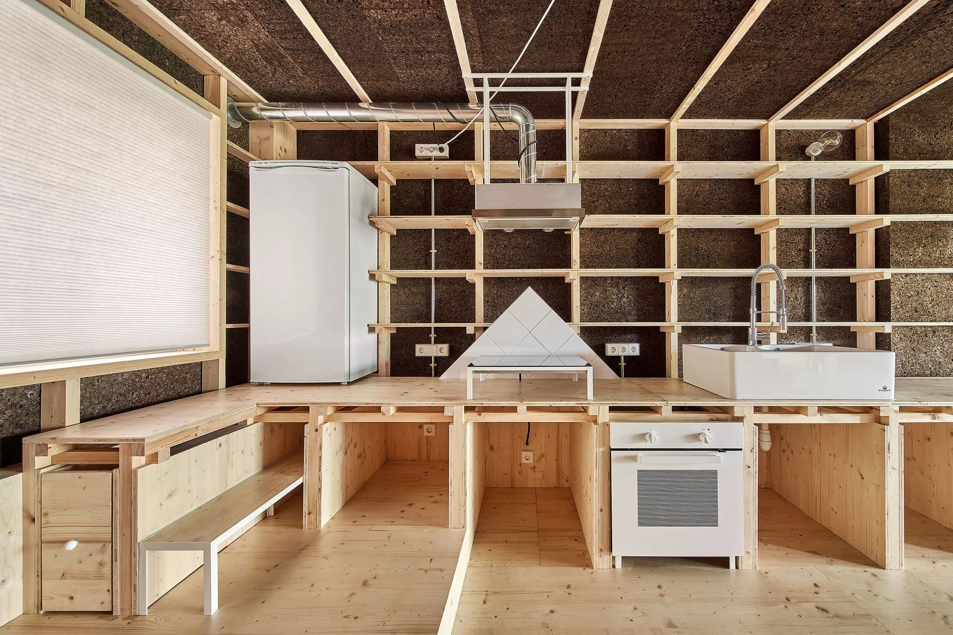 A minimalist kitchen features raw wood framing and dark cork walls with white appliances, a farmhouse sink, and open timber shelves.