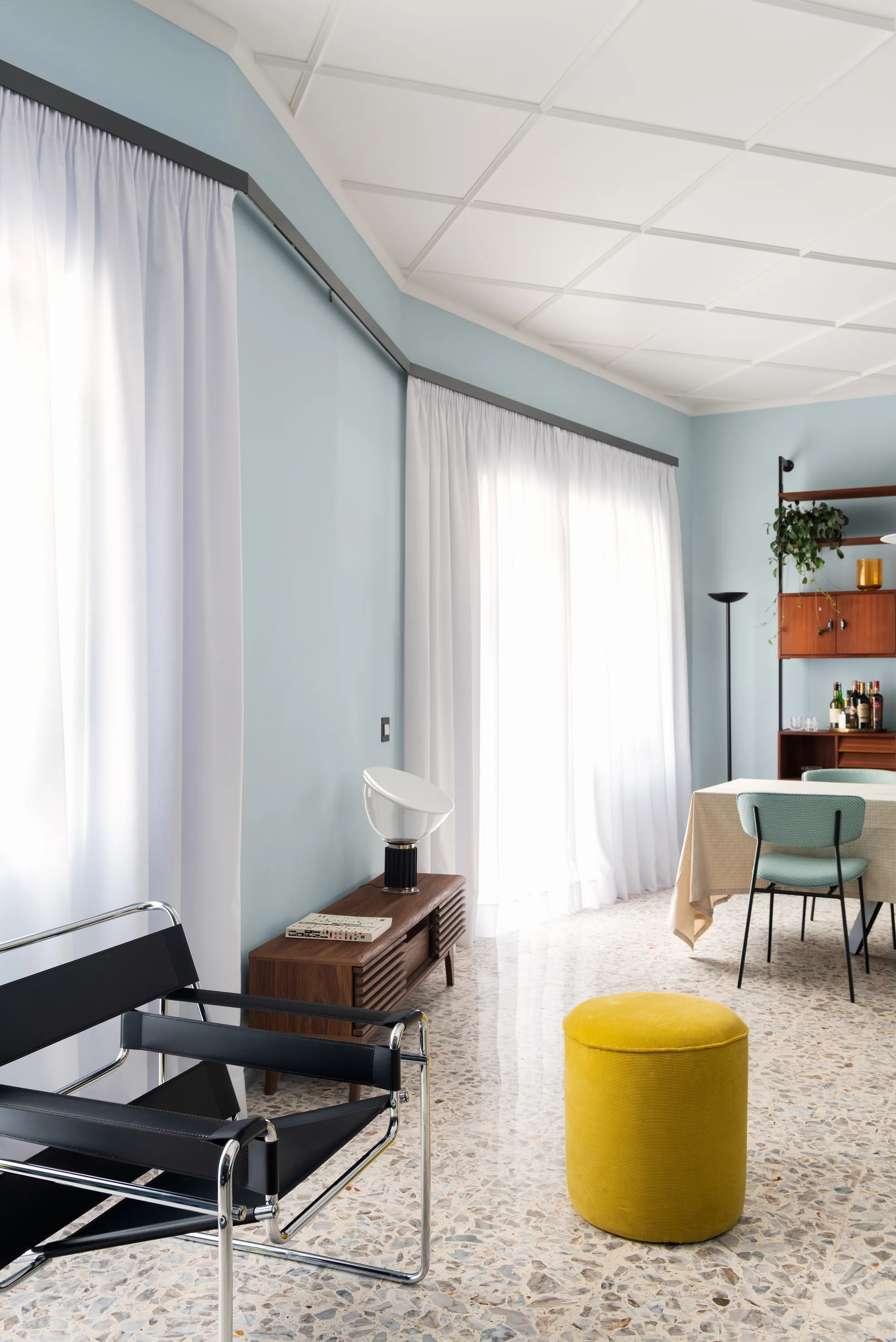 A bright dining space featuring terrazzo tile floors, light blue walls, white sheer curtains, a black lounge chair, and a yellow cylindrical ottoman.