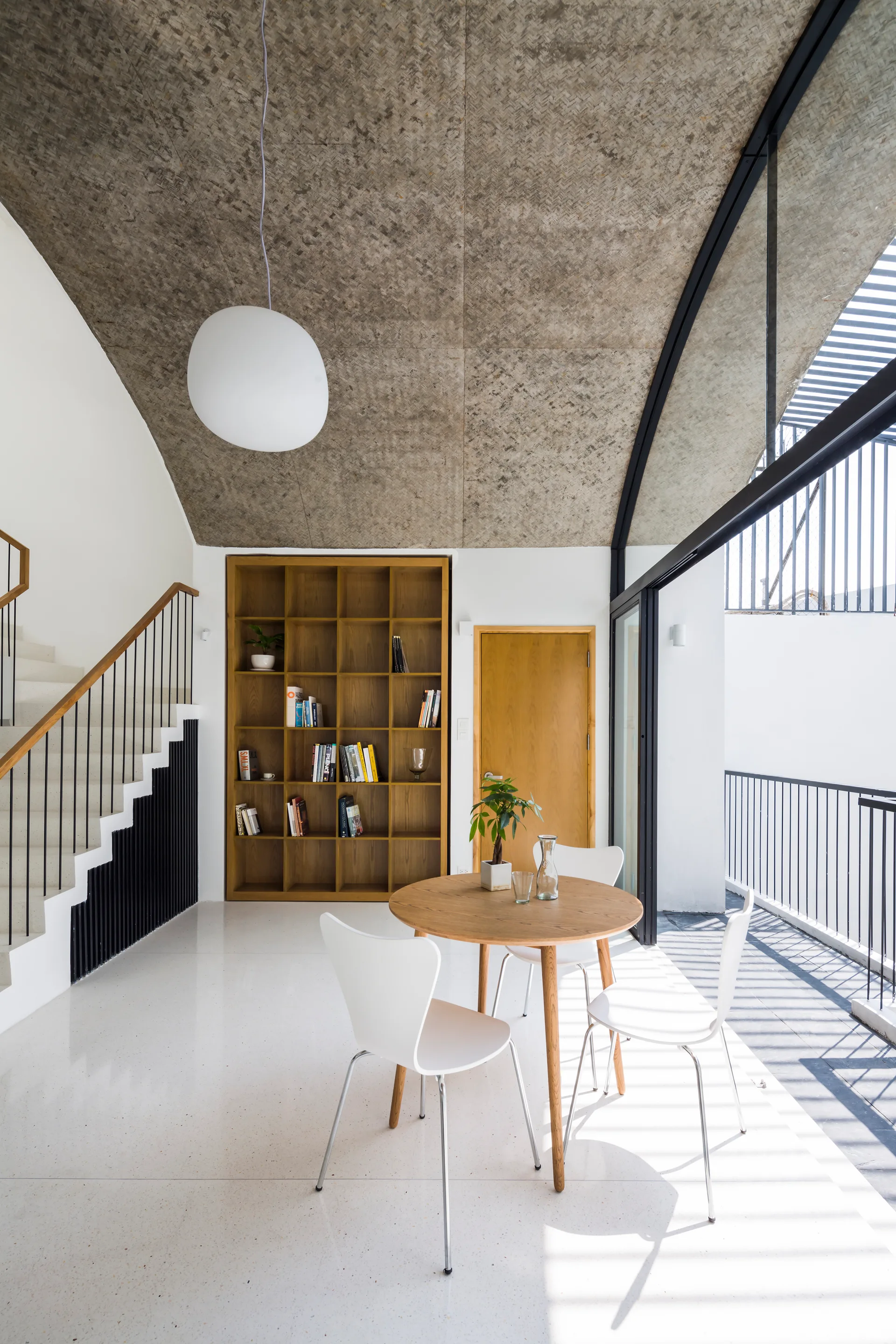 A circular wooden dining table and white chairs sit under a curved woven ceiling next to a built-in bookshelf and white staircase.