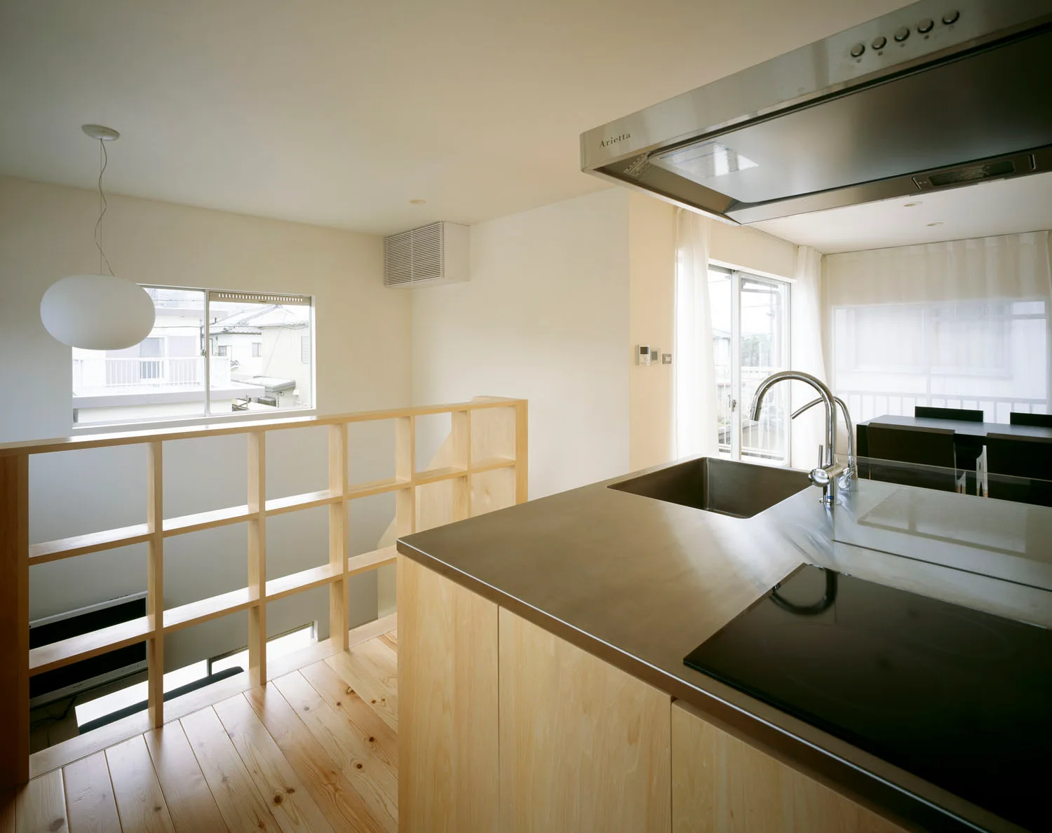 A minimalist kitchen features a stainless steel island with a sink and cooktop, light wood flooring, and an open grid wood railing partition.