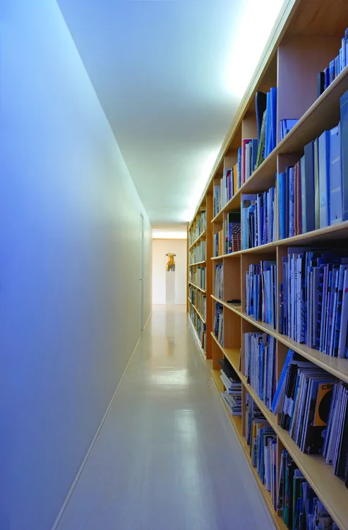 A long hallway featuring custom light wood built-in bookshelves filled with books on one side and a plain wall with linear cove lighting.