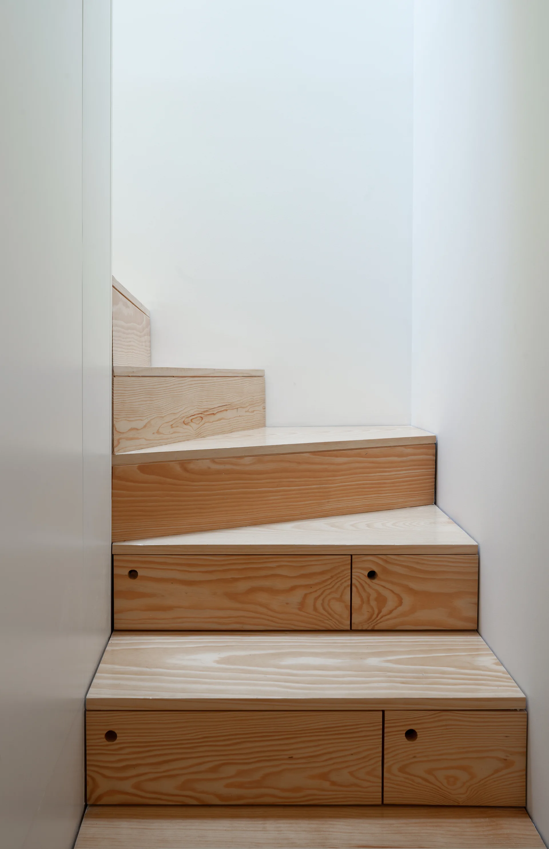A narrow wooden staircase with steps that double as storage drawers, set against smooth white walls.
