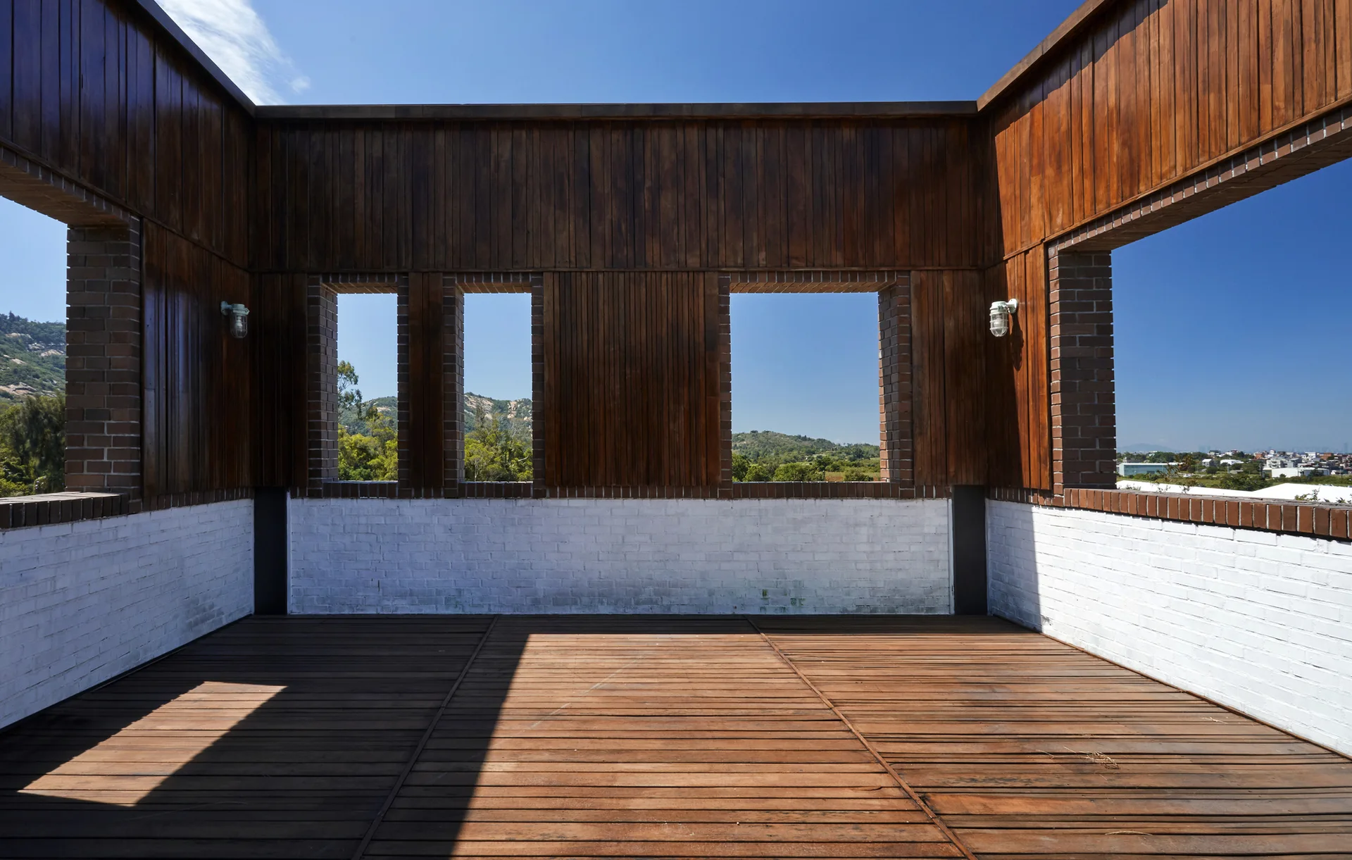 An open-air wooden terrace features white-painted brick walls, vertical wood cladding, and rectangular window cutouts looking out at green hills.