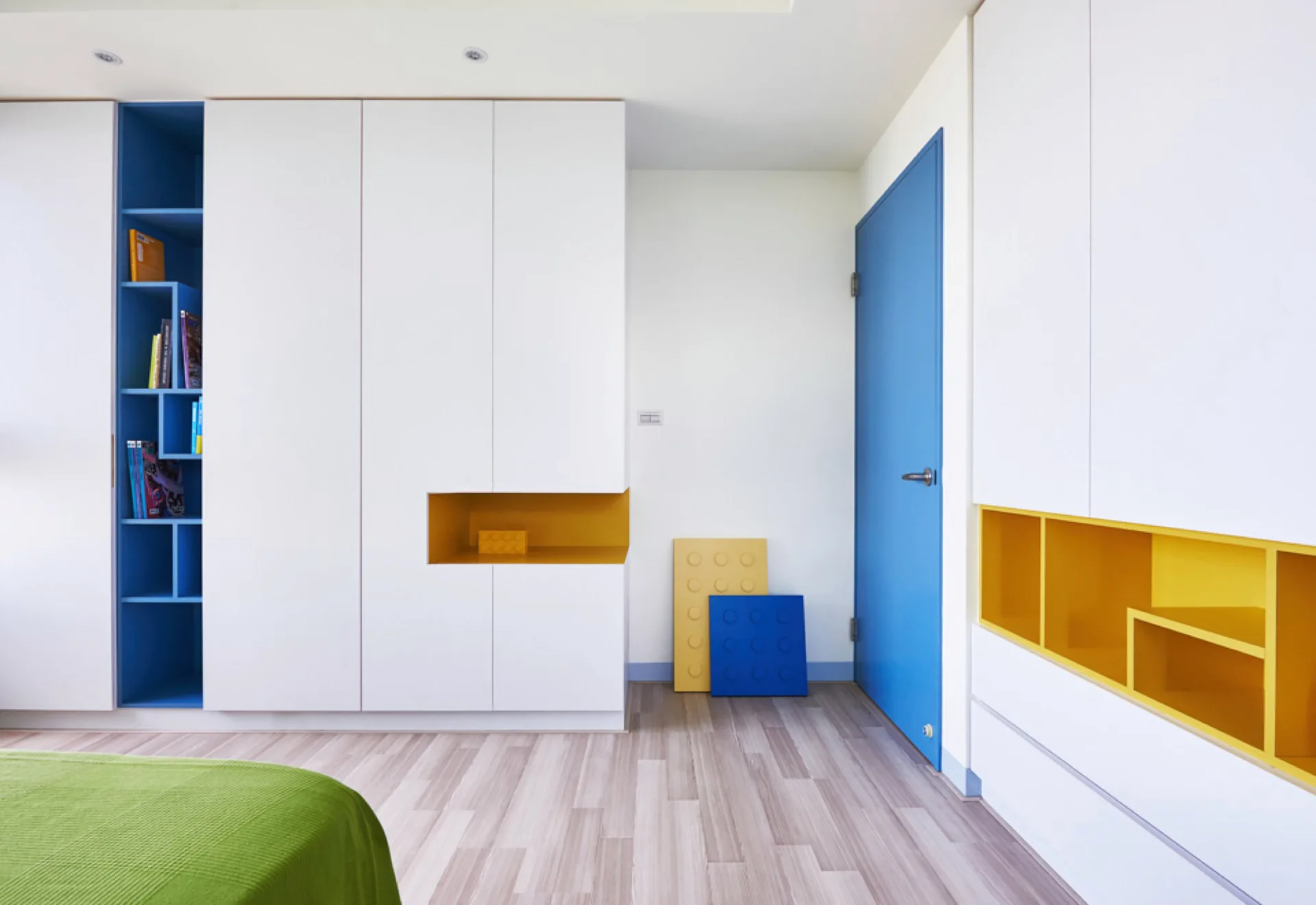 A minimalist children's bedroom features floor-to-ceiling white wardrobes with recessed blue and yellow storage niches and a matching blue door.