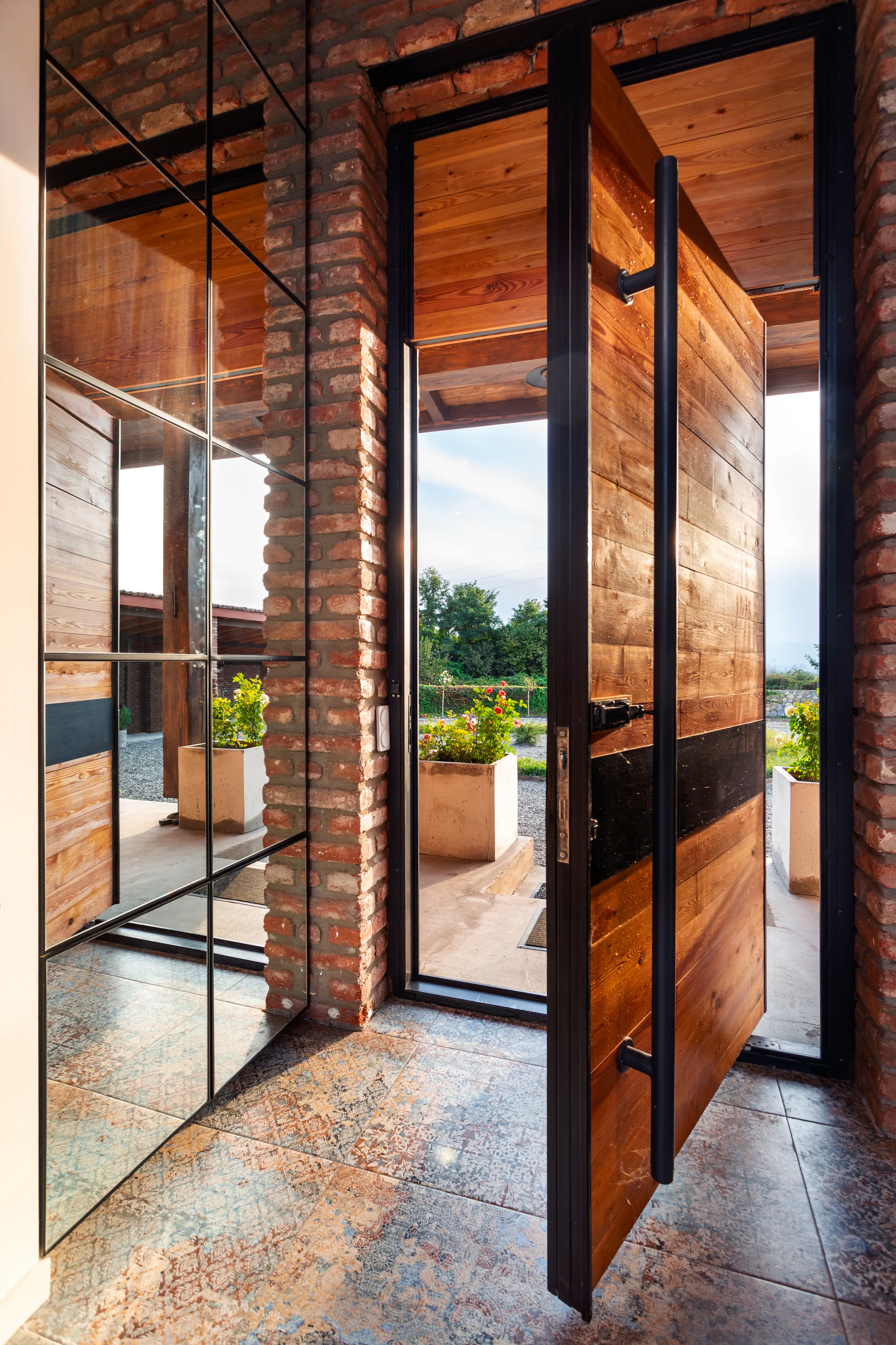 An entryway features a large pivoting wooden door, exposed brick walls, patterned floor tiles, and a grid mirror wall reflecting the space.