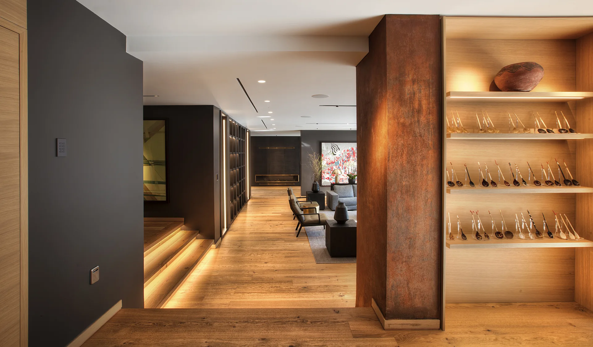 A modern hallway with wood flooring features integrated step lighting, a rust-toned pillar, and built-in wooden display shelving.