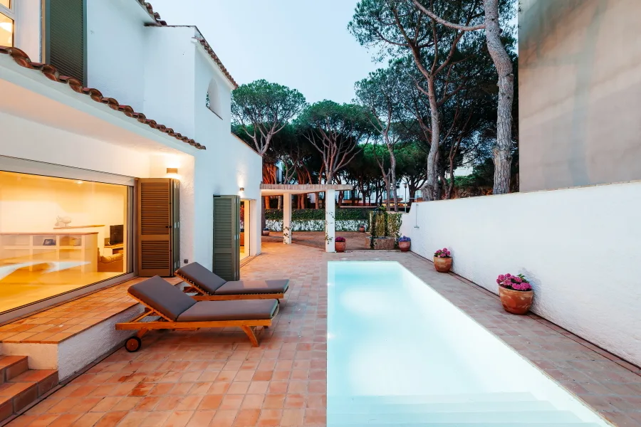 A Mediterranean patio featuring a narrow swimming pool, terracotta tile flooring, wooden lounge chairs, and white stucco walls with green shutters.