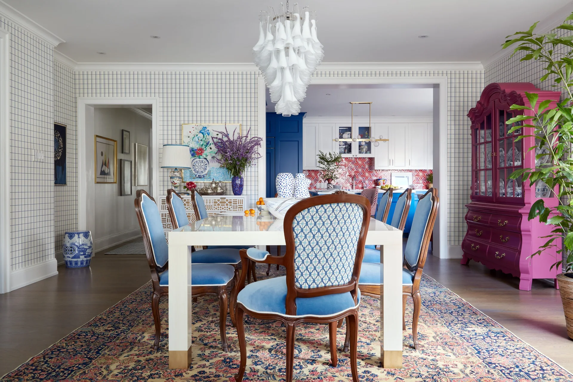 A white dining table surrounded by blue velvet chairs stands on a floral rug in a room with grid wallpaper and a large magenta cabinet.