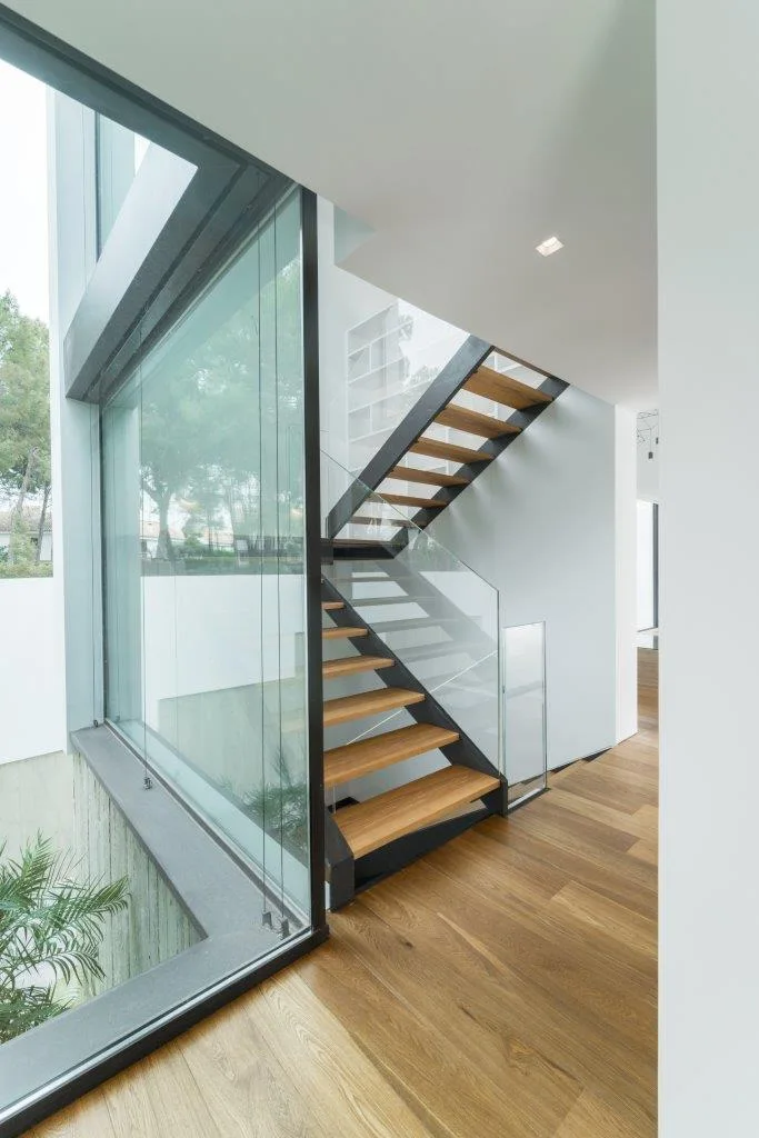 A modern open-riser wood staircase with glass railings sits next to a large floor-to-ceiling glass wall showing an outdoor sunken garden.