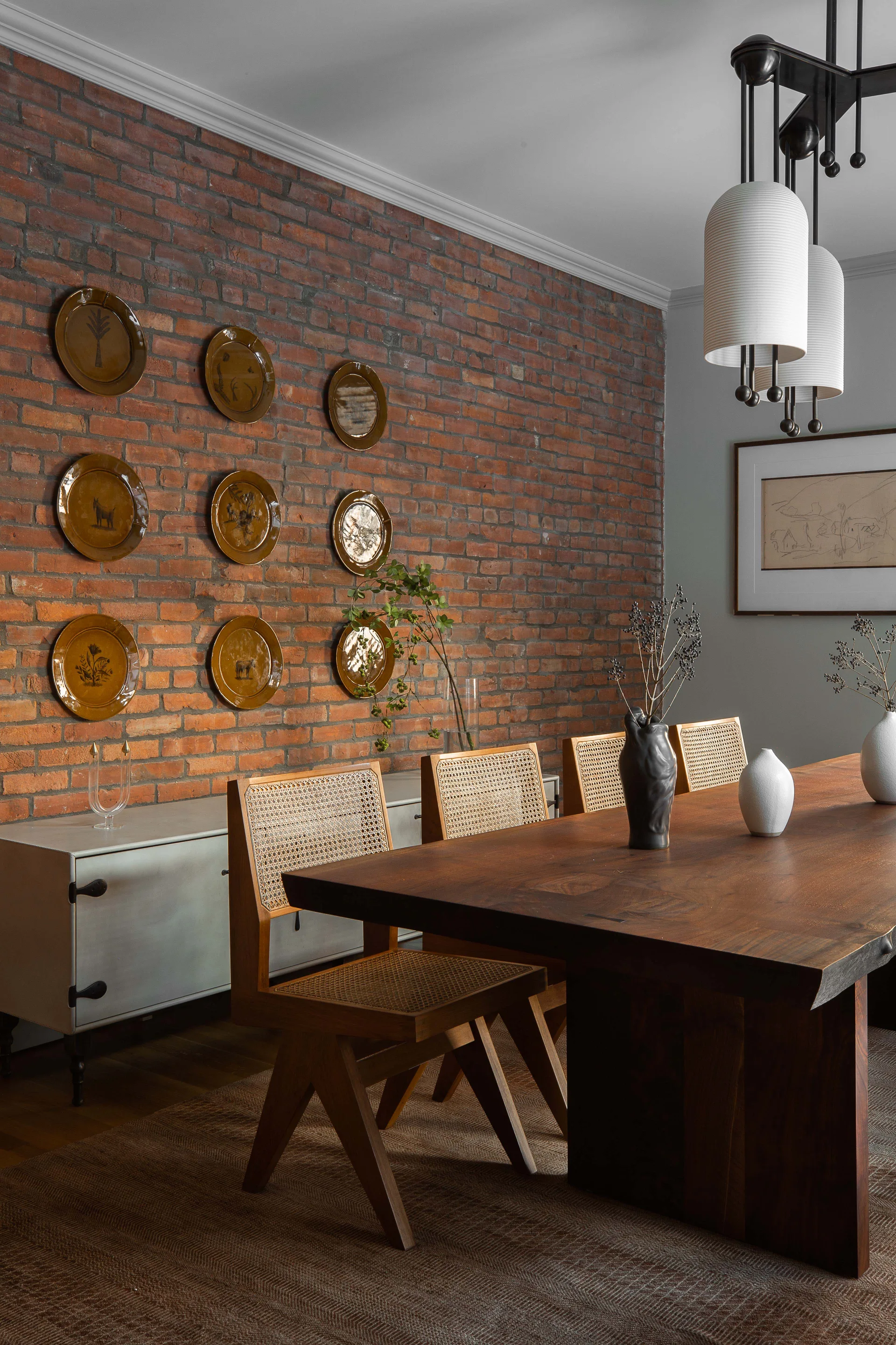 A rustic dining room features a solid wood table, cane-back chairs, and an exposed brick wall decorated with a grid of brown ceramic plates.