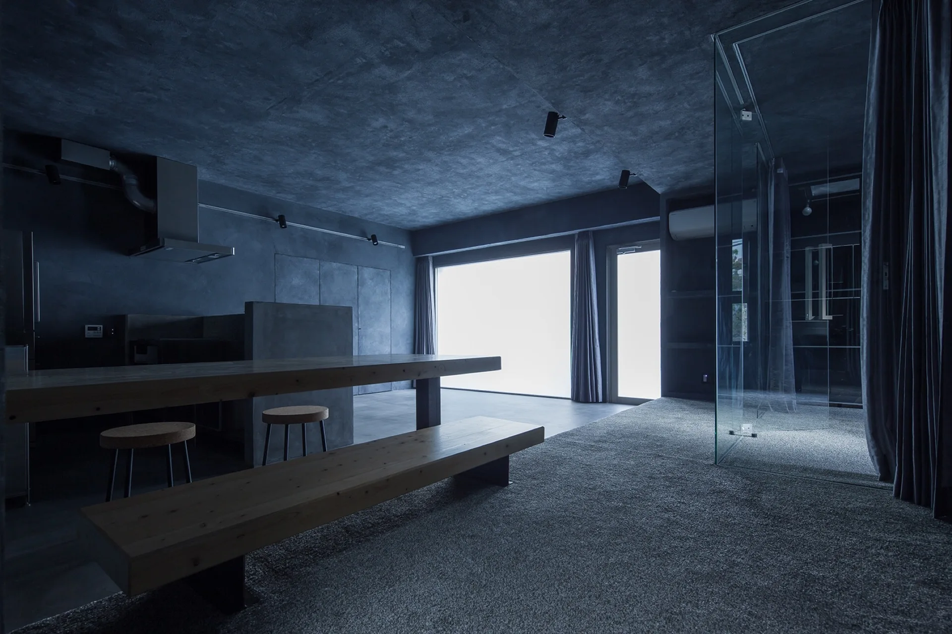 A minimalist dark concrete room features a long wooden table and bench, round stools, and a glass partition near a bright window.